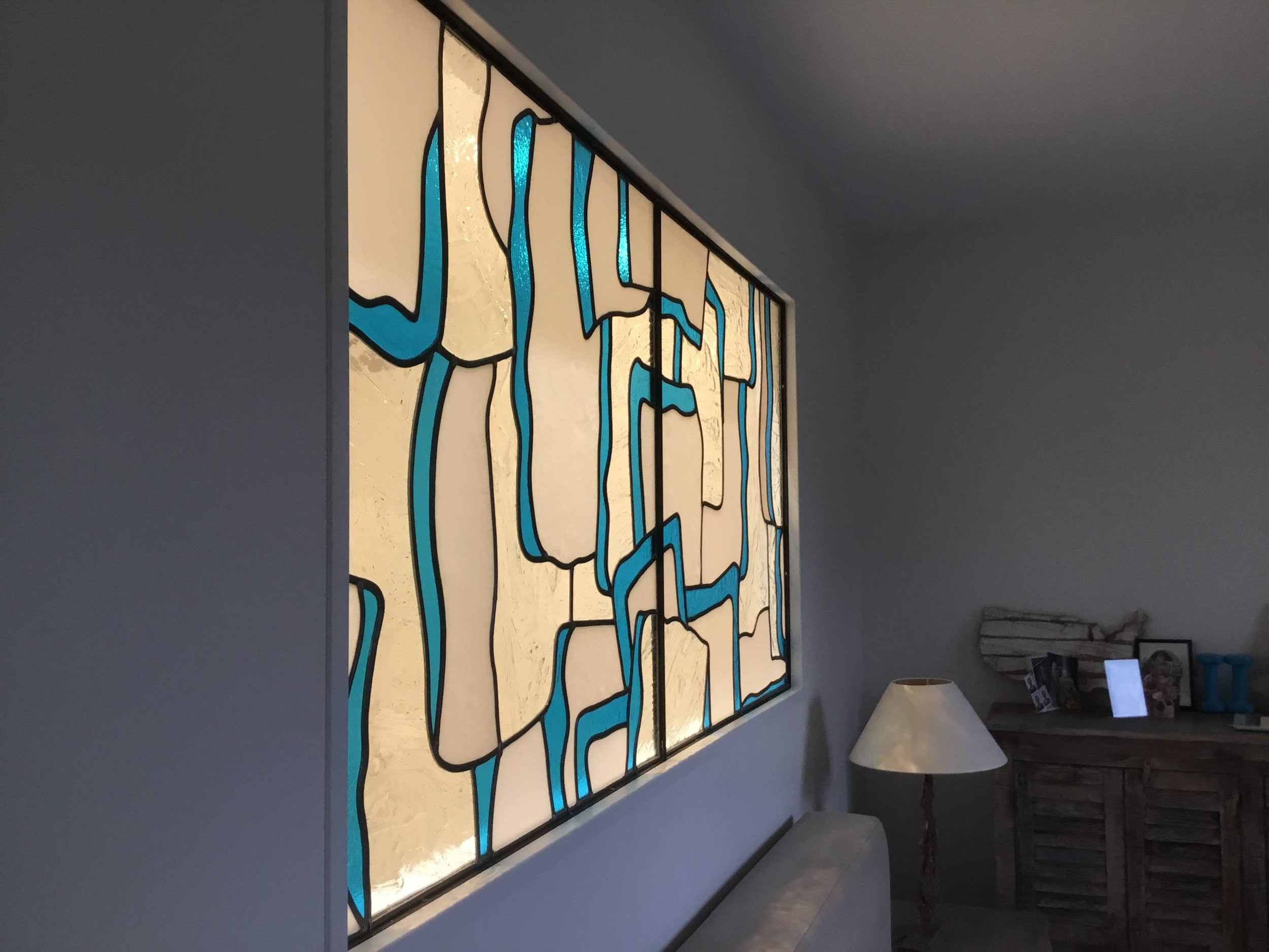 Vitrail au plomb aux motifs contemporains bleu ciel et blanc séparant une chambre et une salle de bain, vu depuis la chambre