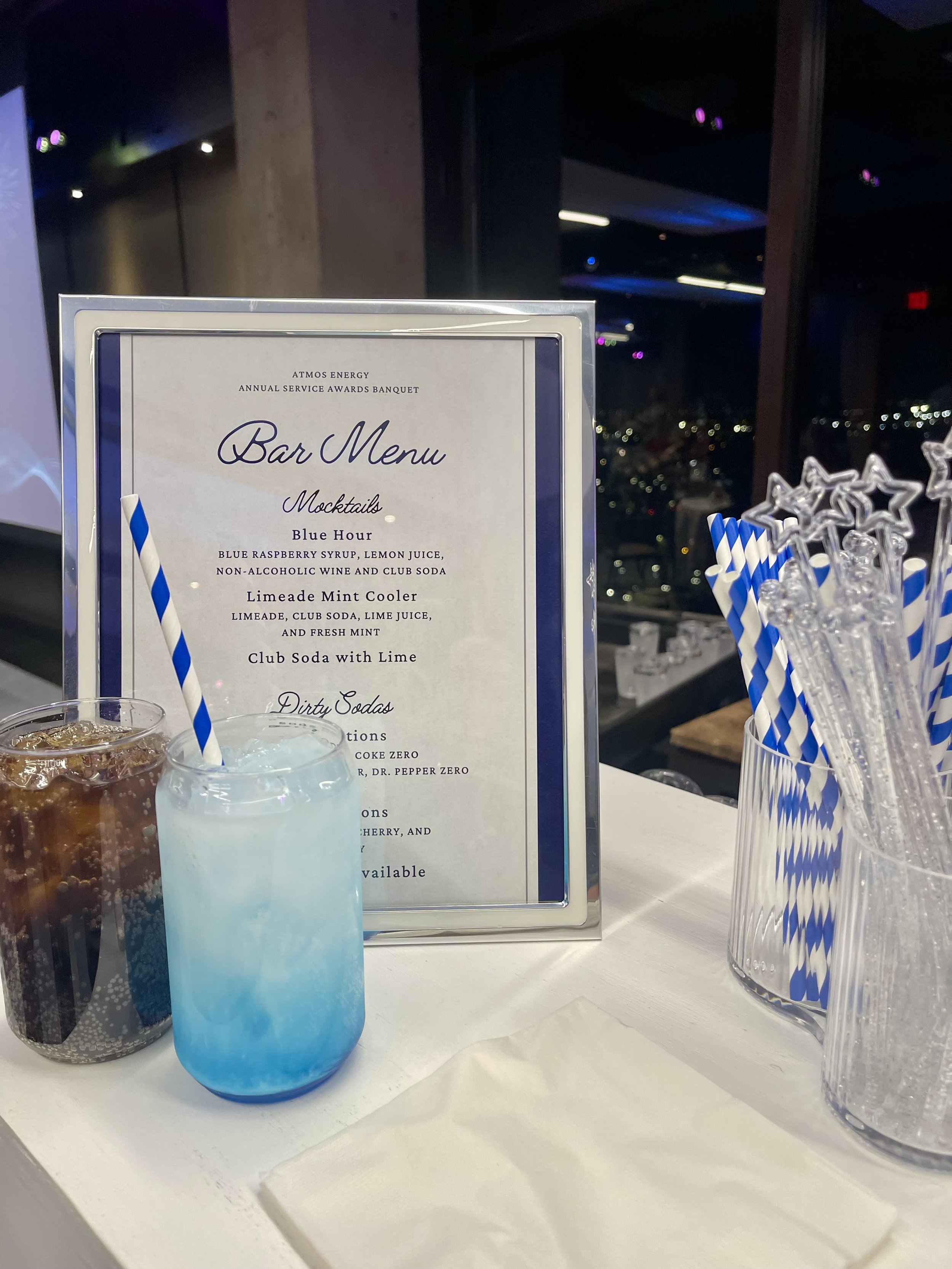 Blue mocktail with striped straw beside an Atmos Energy annual service awards banquet bar menu, cocktail glasses, and decorative straws.