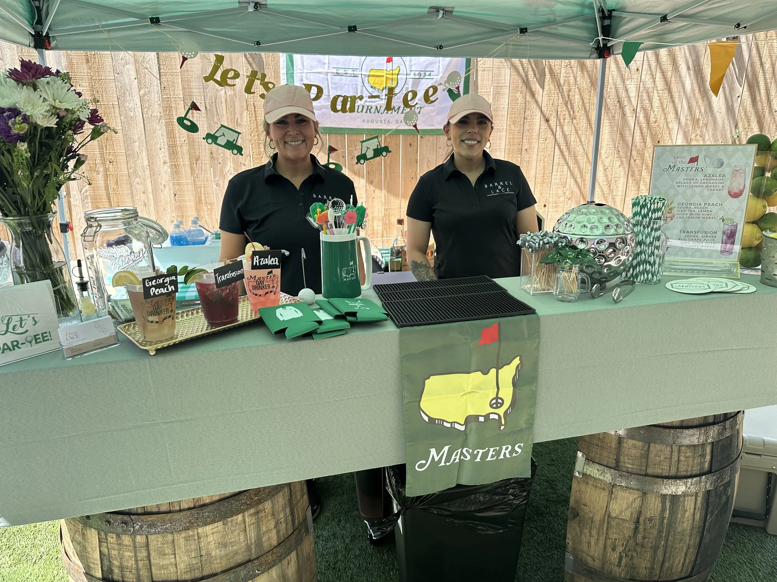Two bartenders serve themed cocktails at a Masters golf tournament drink booth decorated with golf memorabilia and flowers.