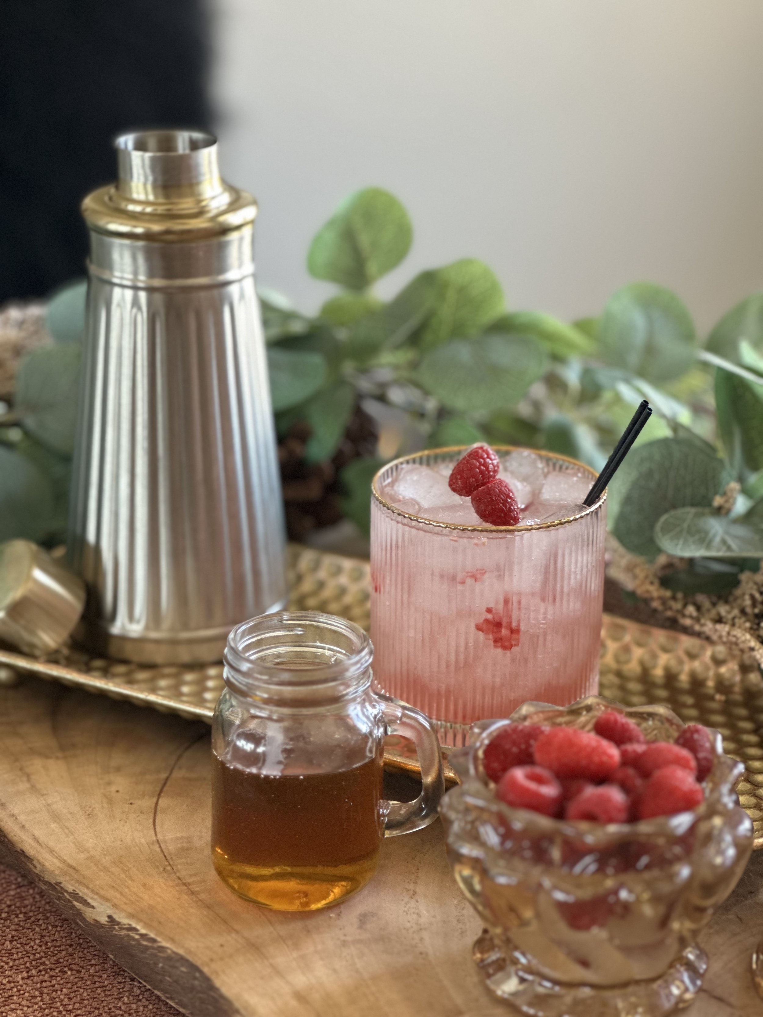 Pink raspberry cocktail with ice and black straw, silver cocktail shaker, brown mixer, and bowl of fresh raspberries on a tray