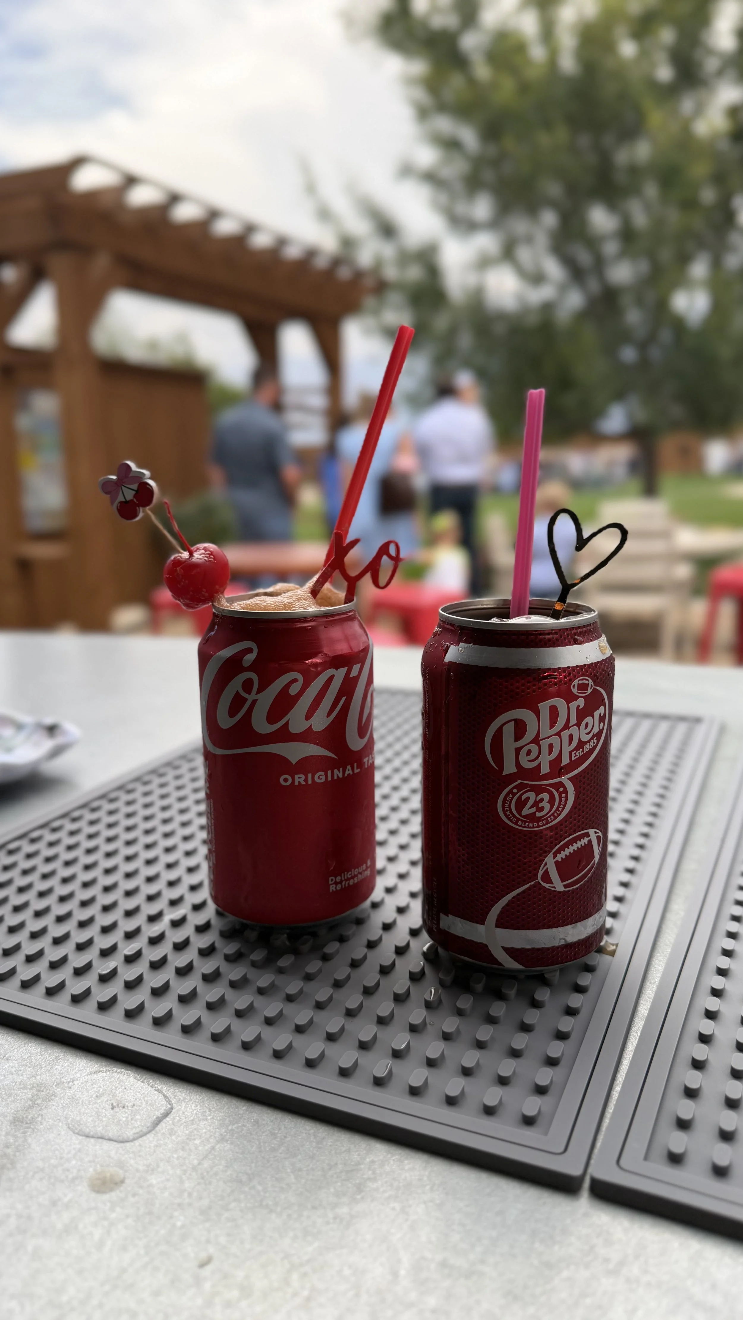 Coca-Cola and Dr Pepper cans with colorful straws and garnishes on an outdoor table as dirty sodas