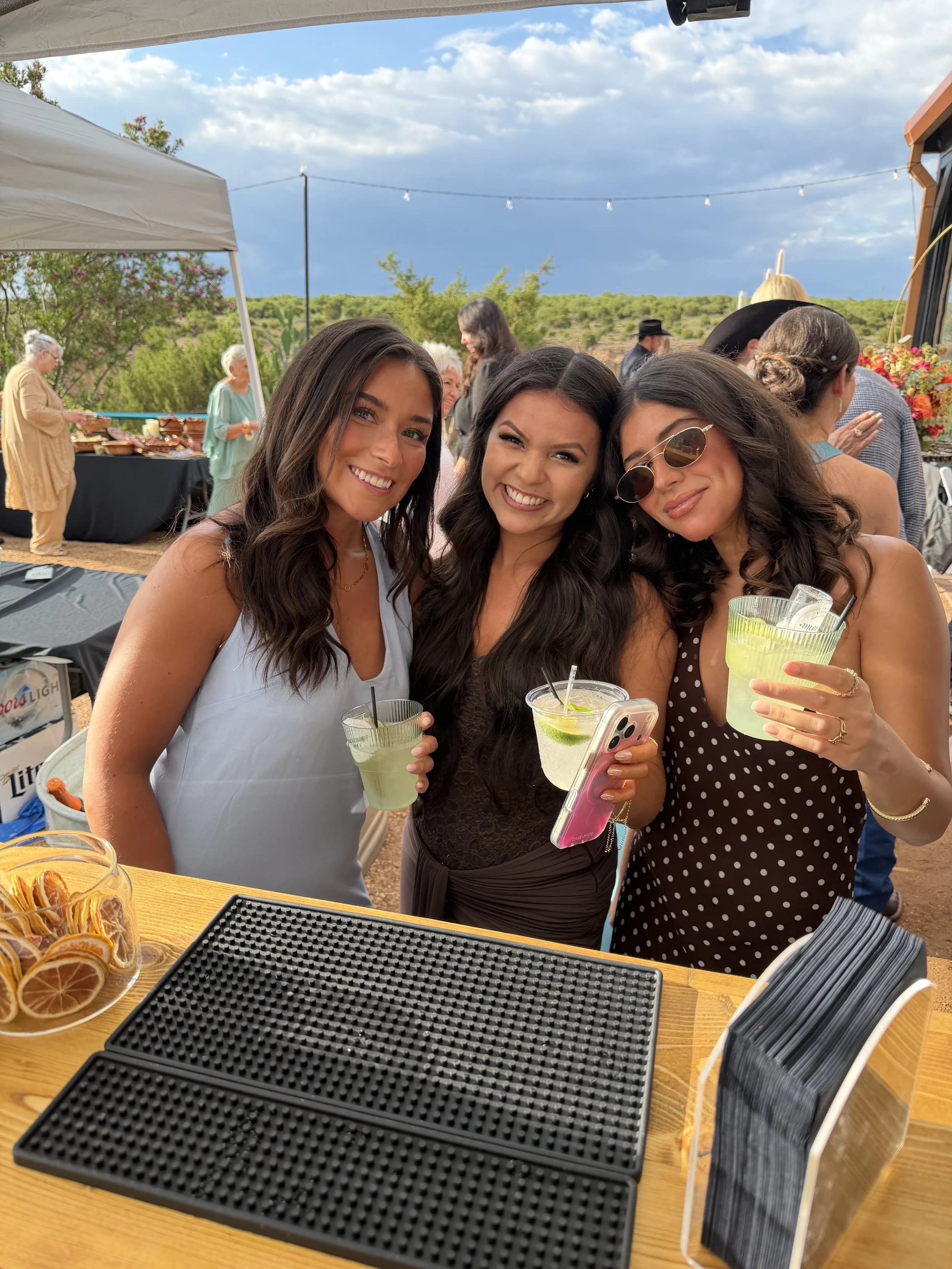 Three women smile for a selfie at an outdoor gathering while holding green cocktails, with string lights, a buffet table, and guests in the background.