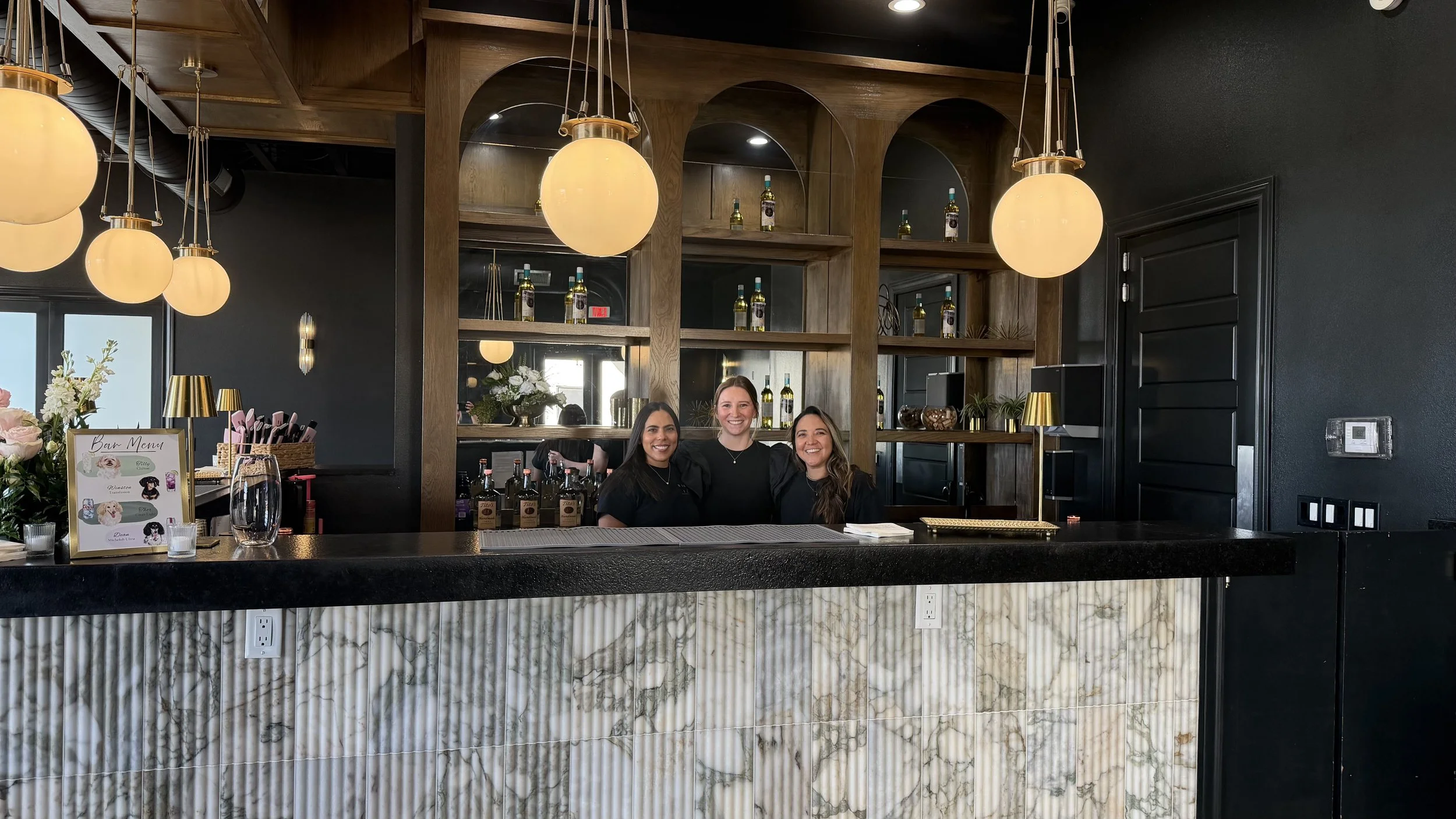 Three smiling bartenders behind a stylish black bar with liquor bottles, mirrors, plants, and globe pendant lights
