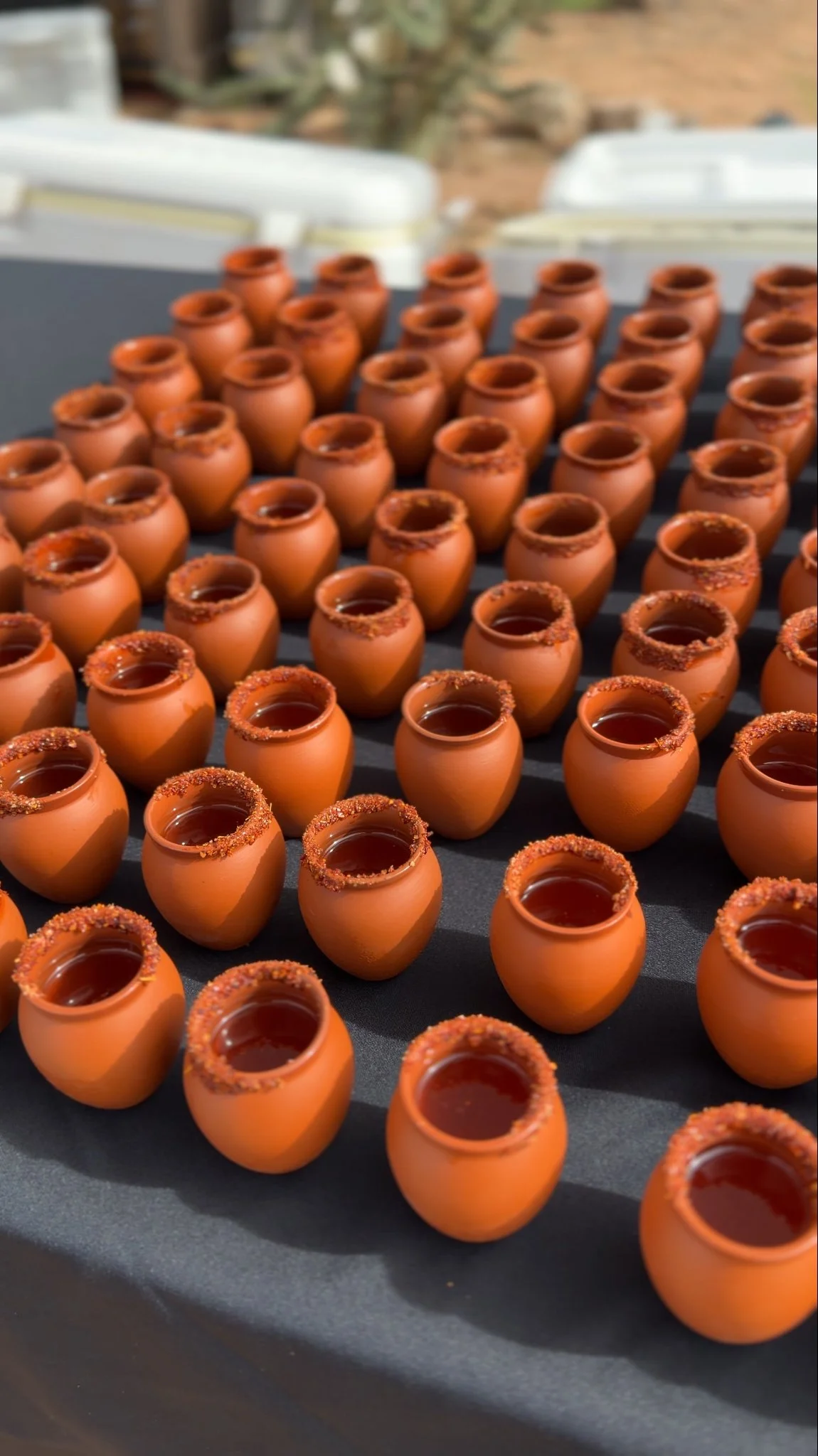 Rows of small terracotta cups filled with a dark red drink, many with chili-salt rims, displayed on a black table.