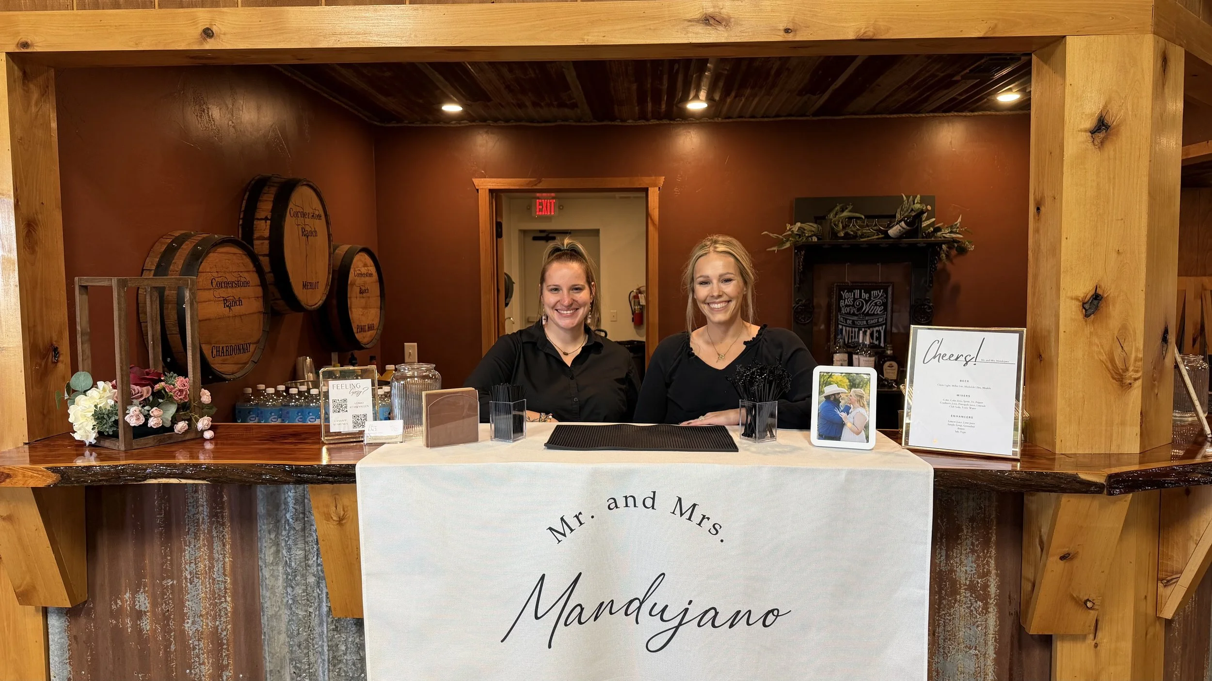 Two smiling bartenders behind a rustic wedding bar with wine barrels, cocktail supplies, and a custom linen sign.