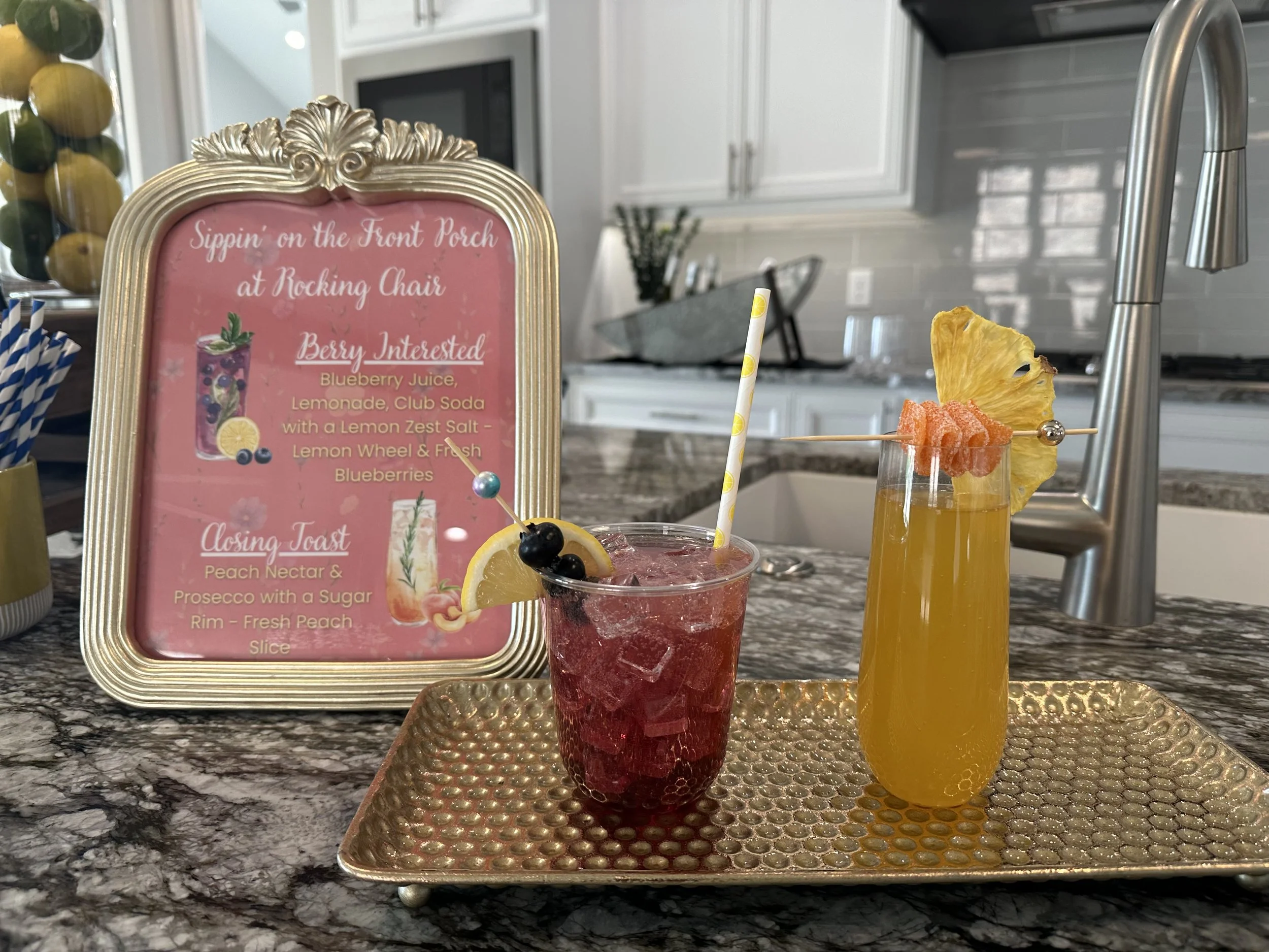 Two colorful fruit cocktails on a tray in a kitchen, garnished with blueberries, lemon, candied citrus, and dried pineapple beside a drink recipe sign.