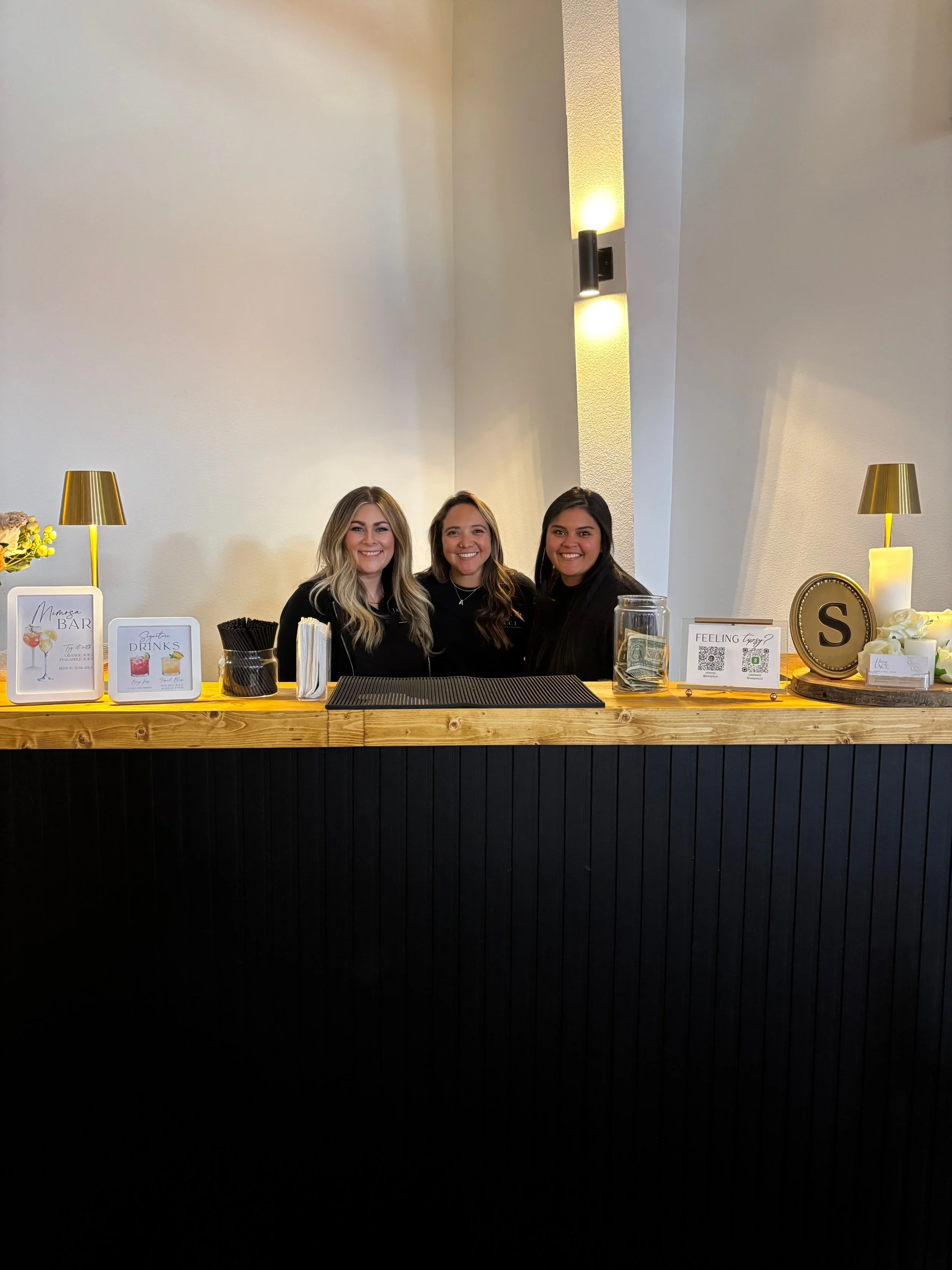 Three smiling women stand behind a decorated event bar with drink menus, straws, napkins, a tip jar, and warm lighting.