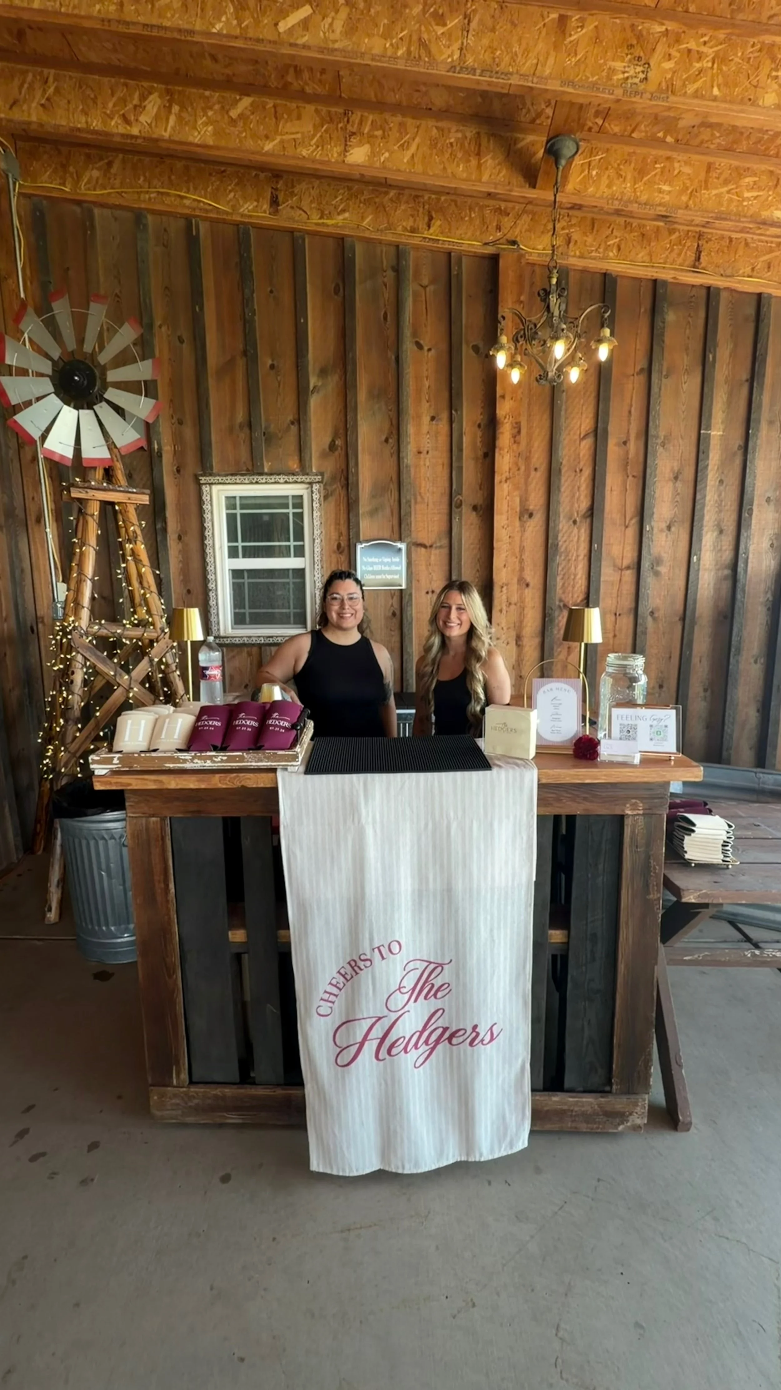 Two women behind a rustic event bar with branded cups, a banner reading “Cheers to the Hedges,” and a decorative windmill.