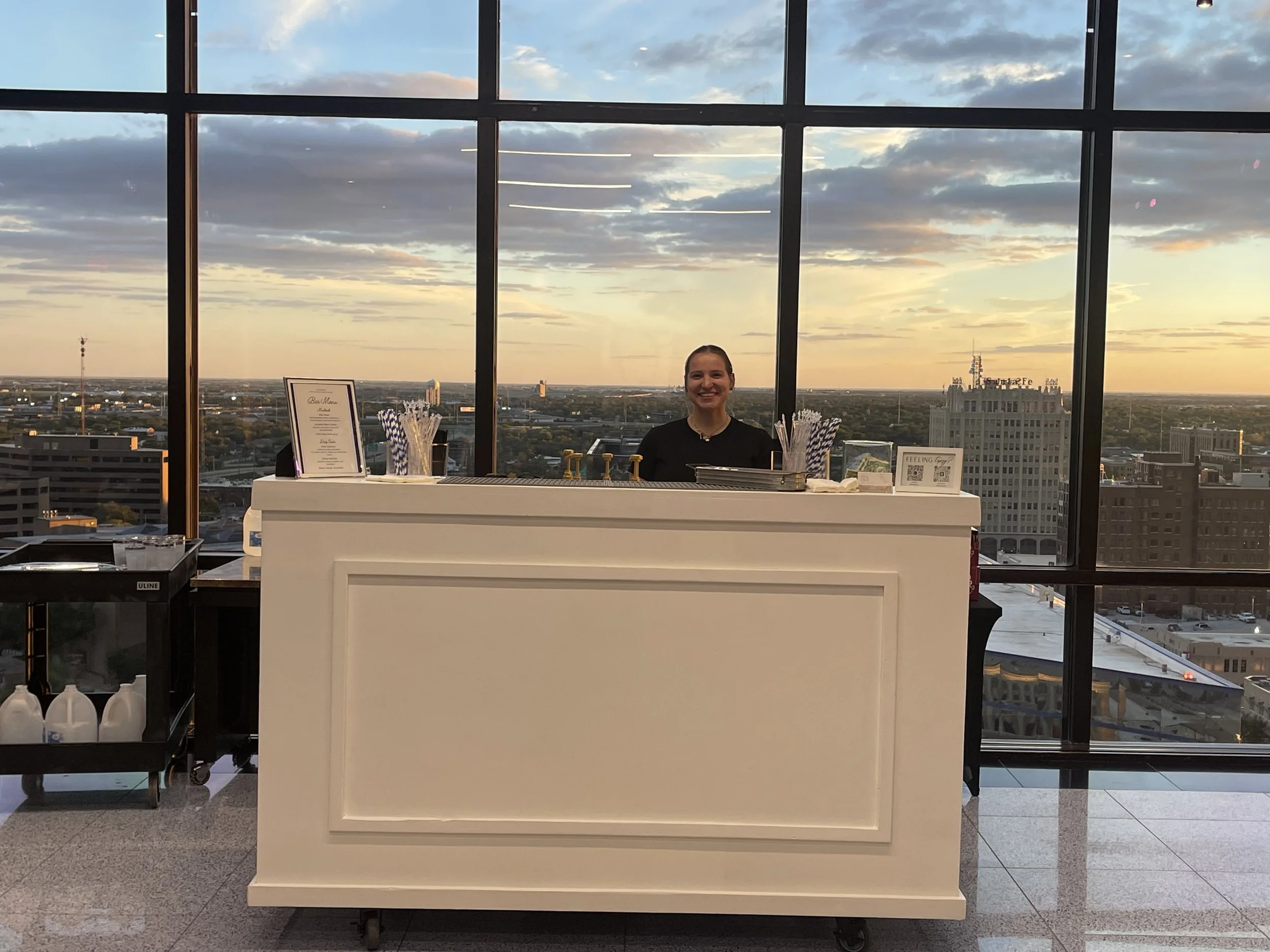 Bartender smiling behind a white bar cart with a city skyline and sunset visible through floor-to-ceiling windows