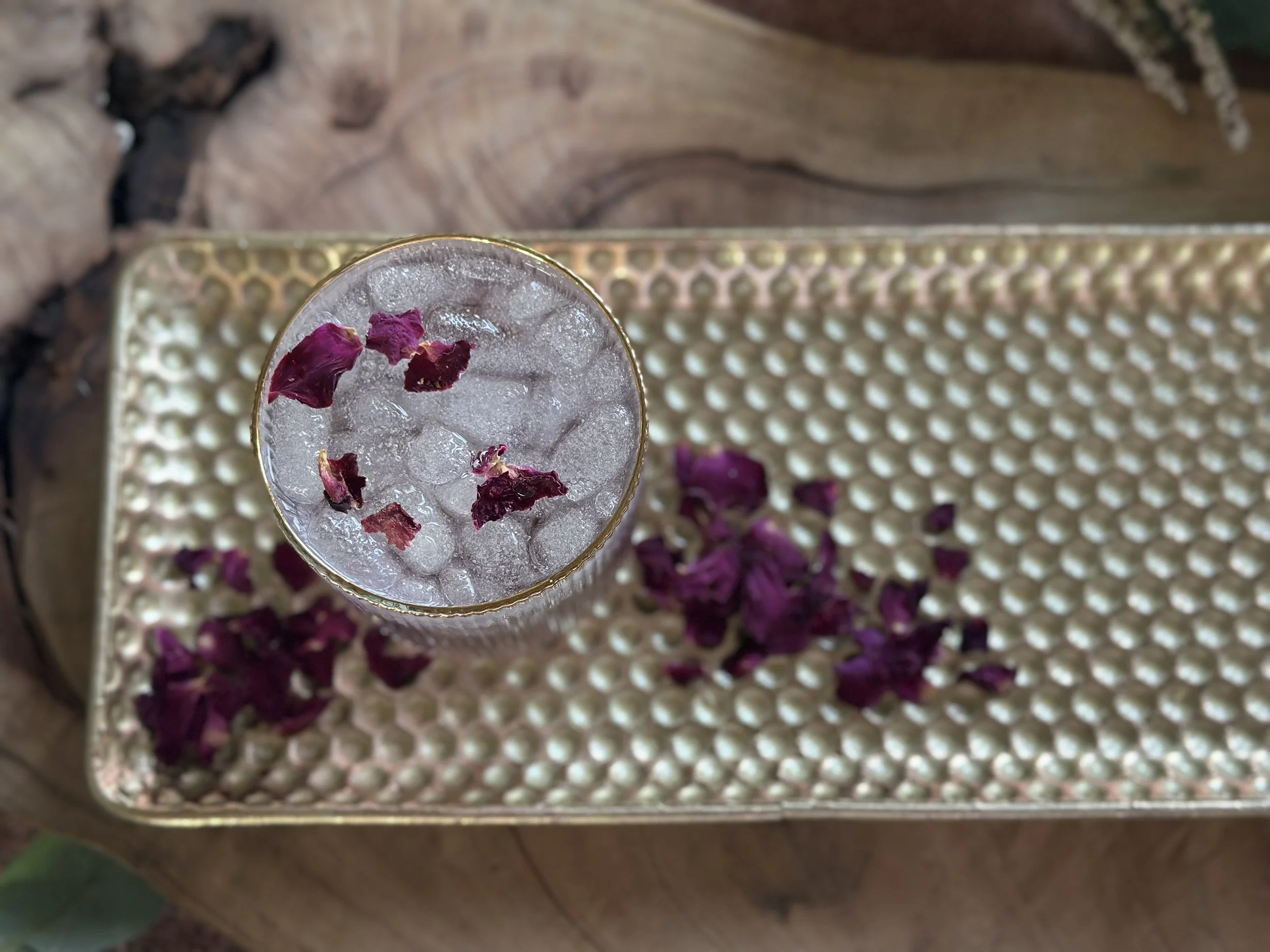 Top-down view of a gold-rimmed glass filled with ice and dried rose petals on a decorative tray