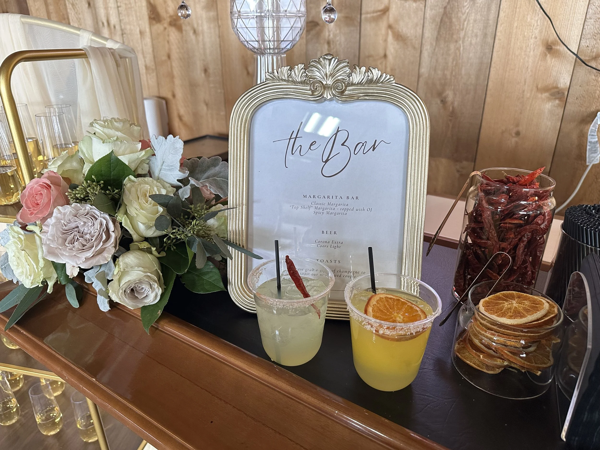 Margarita bar display with framed menu, two colorful cocktails, flowers, dried citrus slices, and dried red chilies