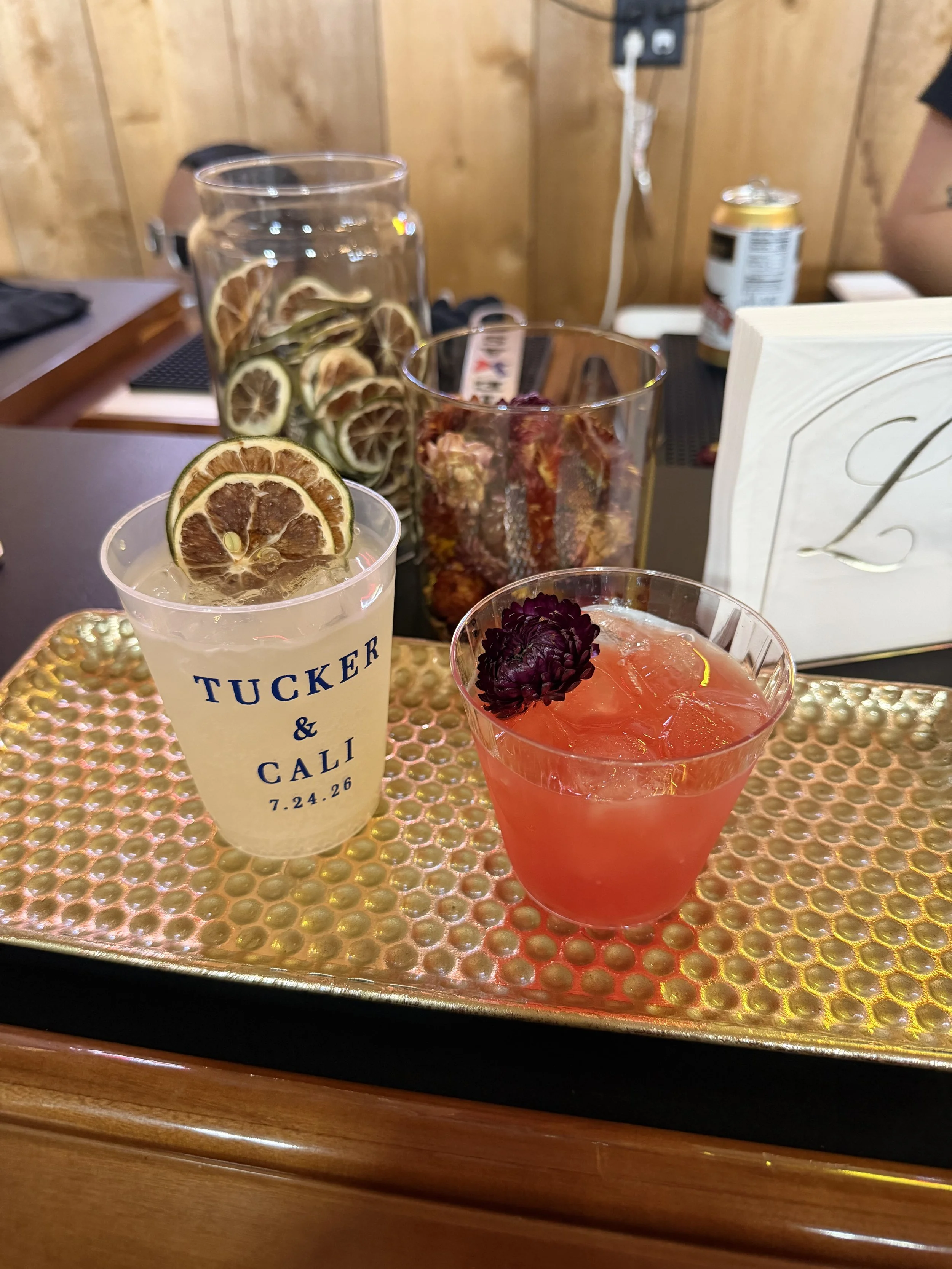 Two colorful cocktails garnished with dried citrus slices and a purple flower on a textured bar tray.