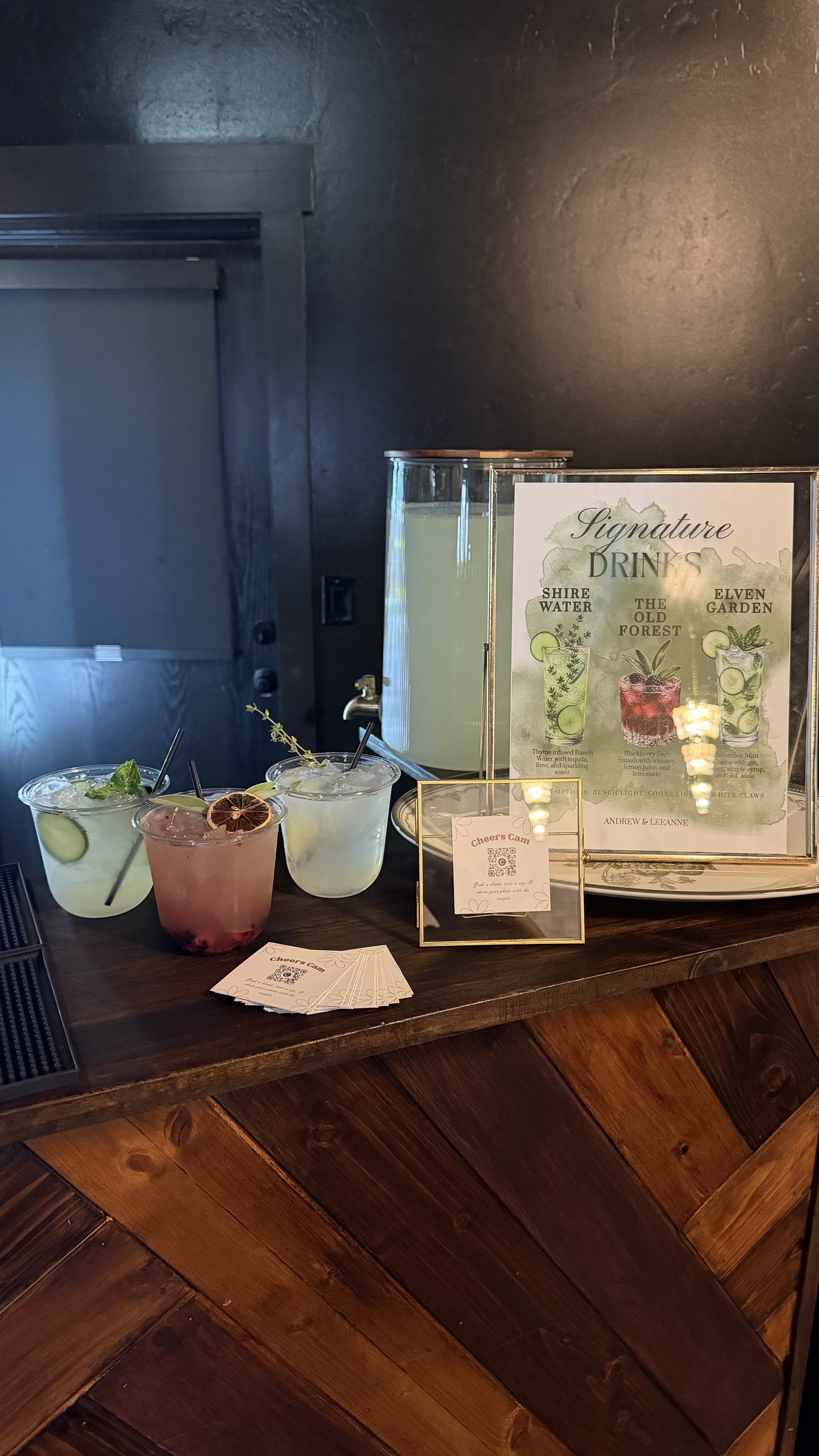 Three colorful cocktails with garnishes beside a framed signature drinks menu and Cheers Cam cards on a rustic wooden bar.