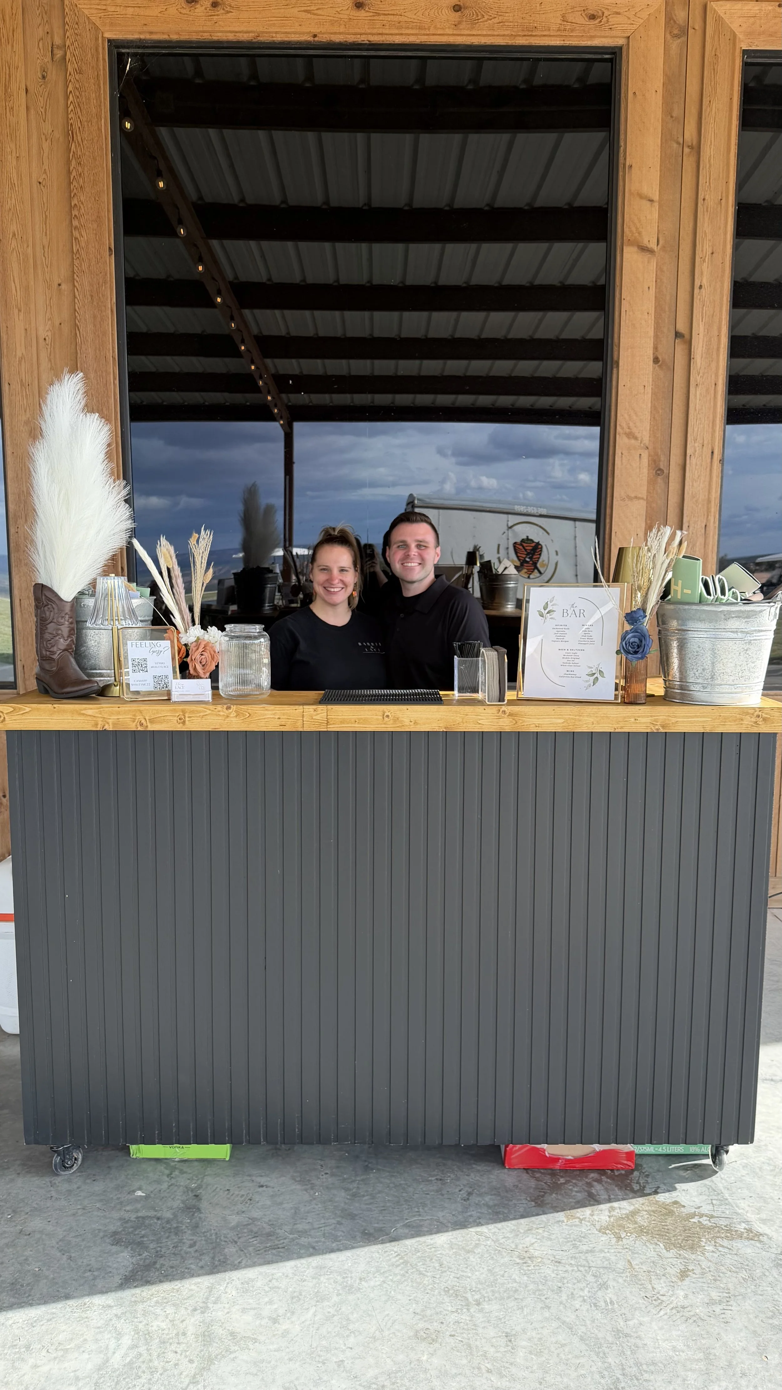 Two bartenders pose behind a rustic wooden bar decorated with flowers, dried grasses, menus, and drinkware.