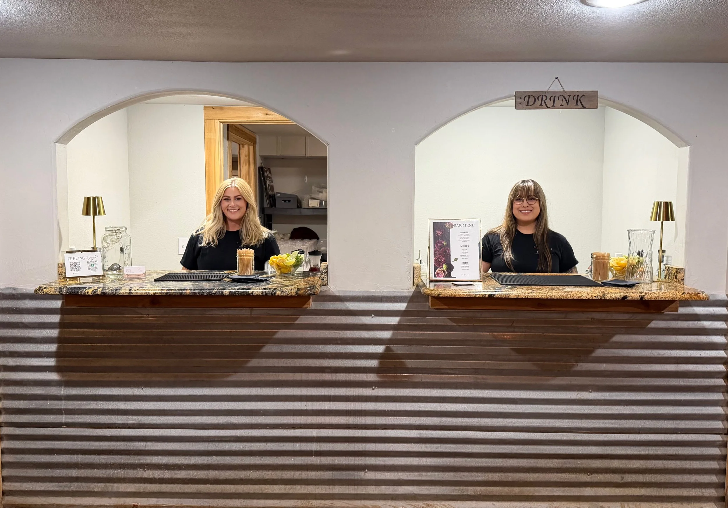 Two bartenders behind a bar counter with drink ingredients, glassware, a bar menu, and a hanging “DRINK” sign