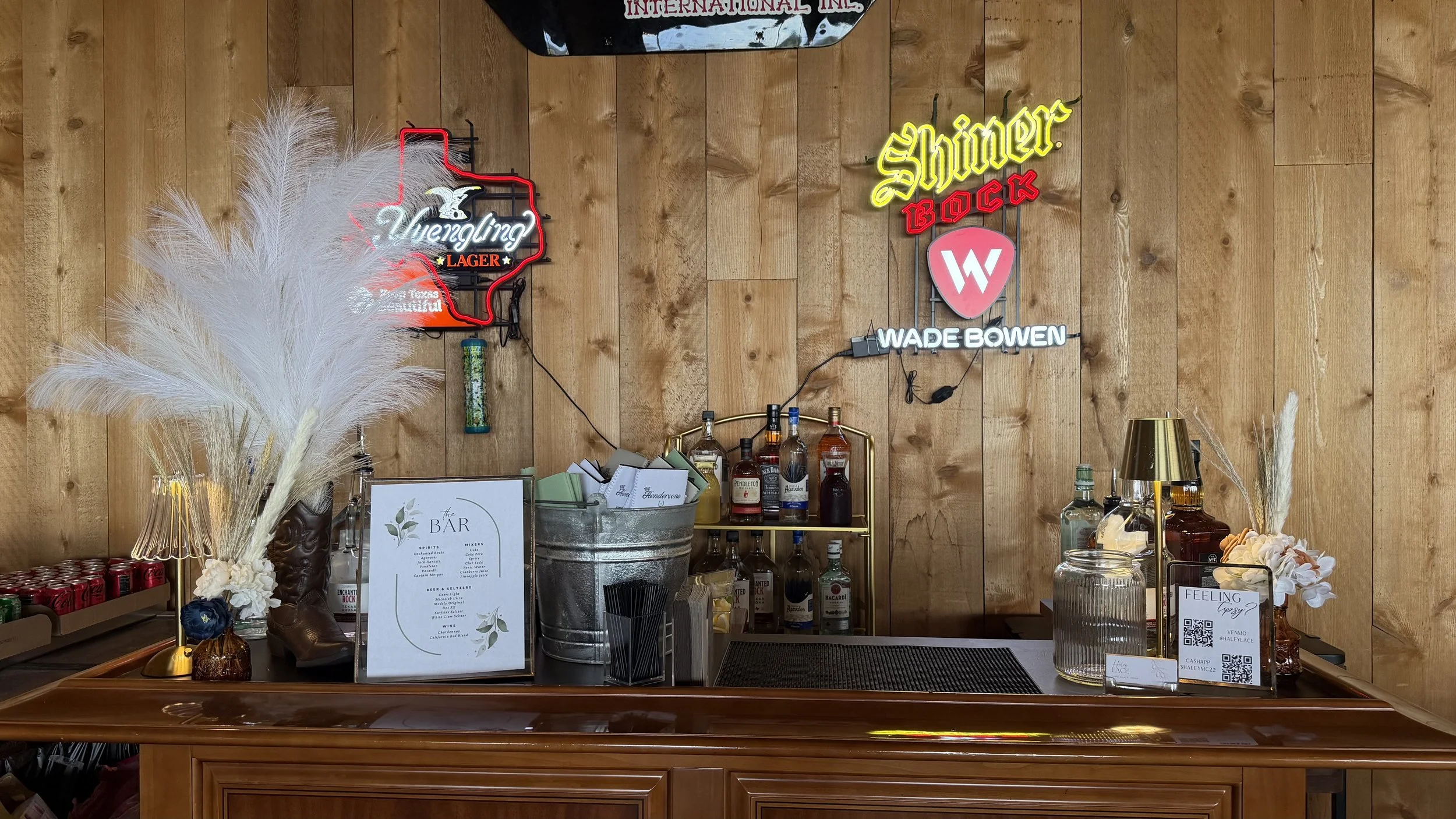 Rustic wood-paneled bar with liquor bottles, cocktail supplies, floral decor, and illuminated Shiner Bock and Weihenstephan neon signs.