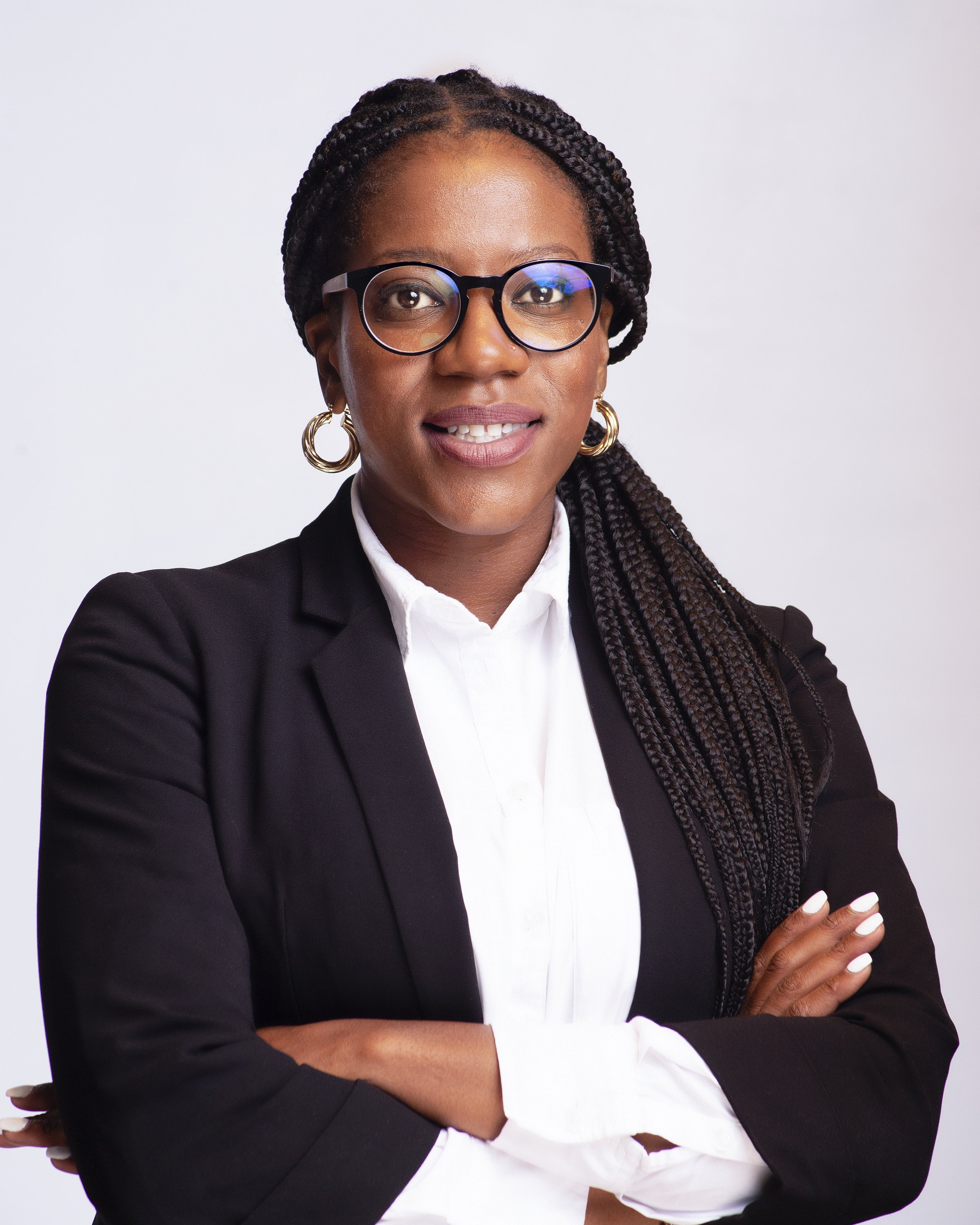 A smiling woman with curly hair, glasses, hoop earrings, and layered necklaces, indoors with a blurred background.