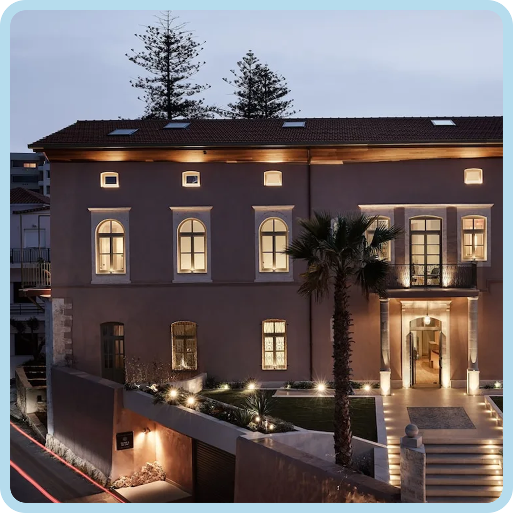A modern multi-story house illuminated at dusk with outdoor lighting, featuring a palm tree in front and a staircase leading to the entrance.