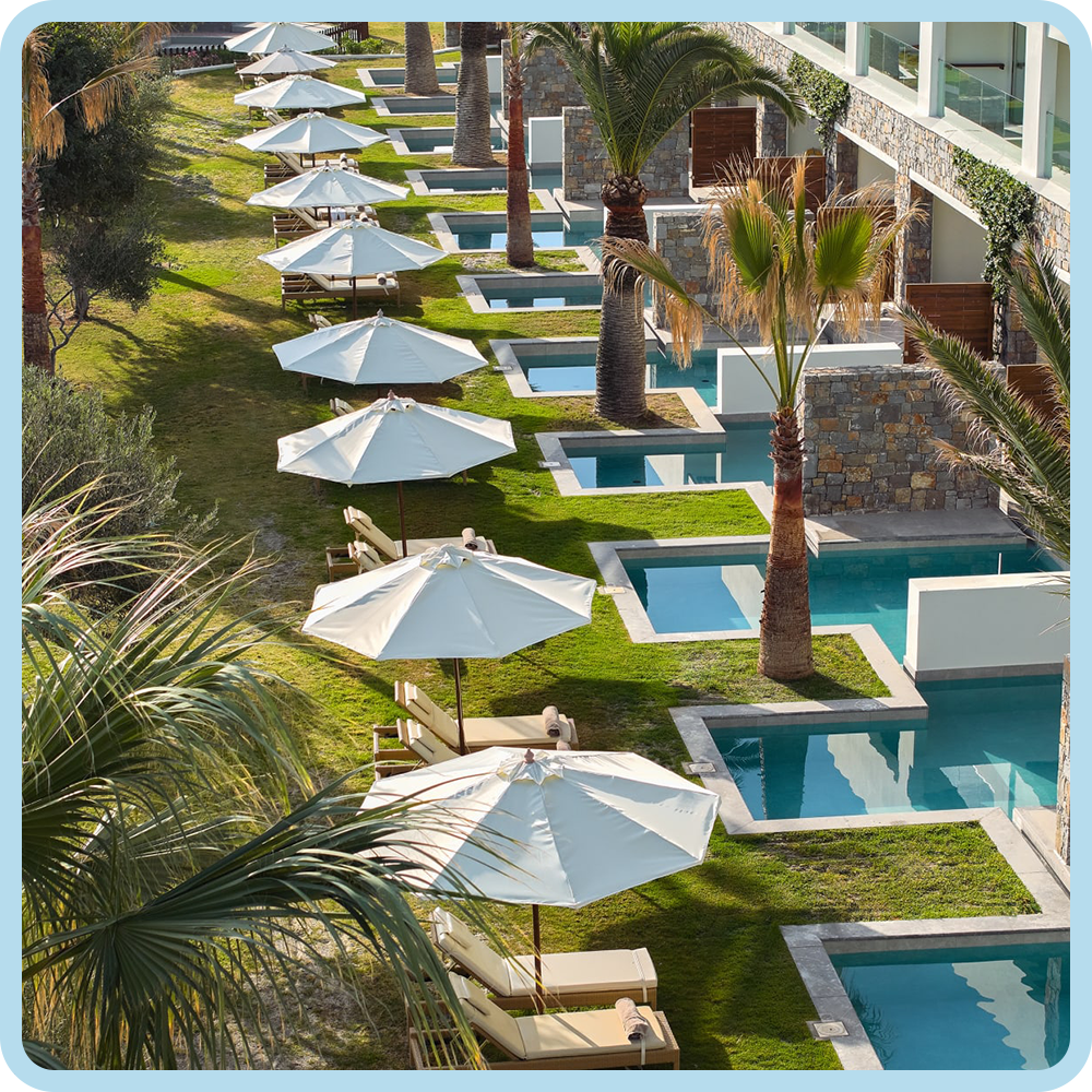 Poolside lounge chairs with white umbrellas alongside a multi-tiered swimming pool surrounded by palm trees and greenery at a resort.
