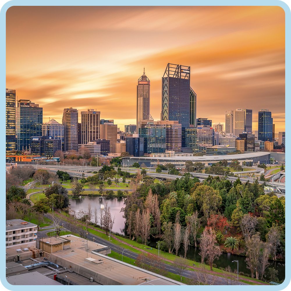 City skyline at sunset with tall buildings and a park with trees and water in the foreground.