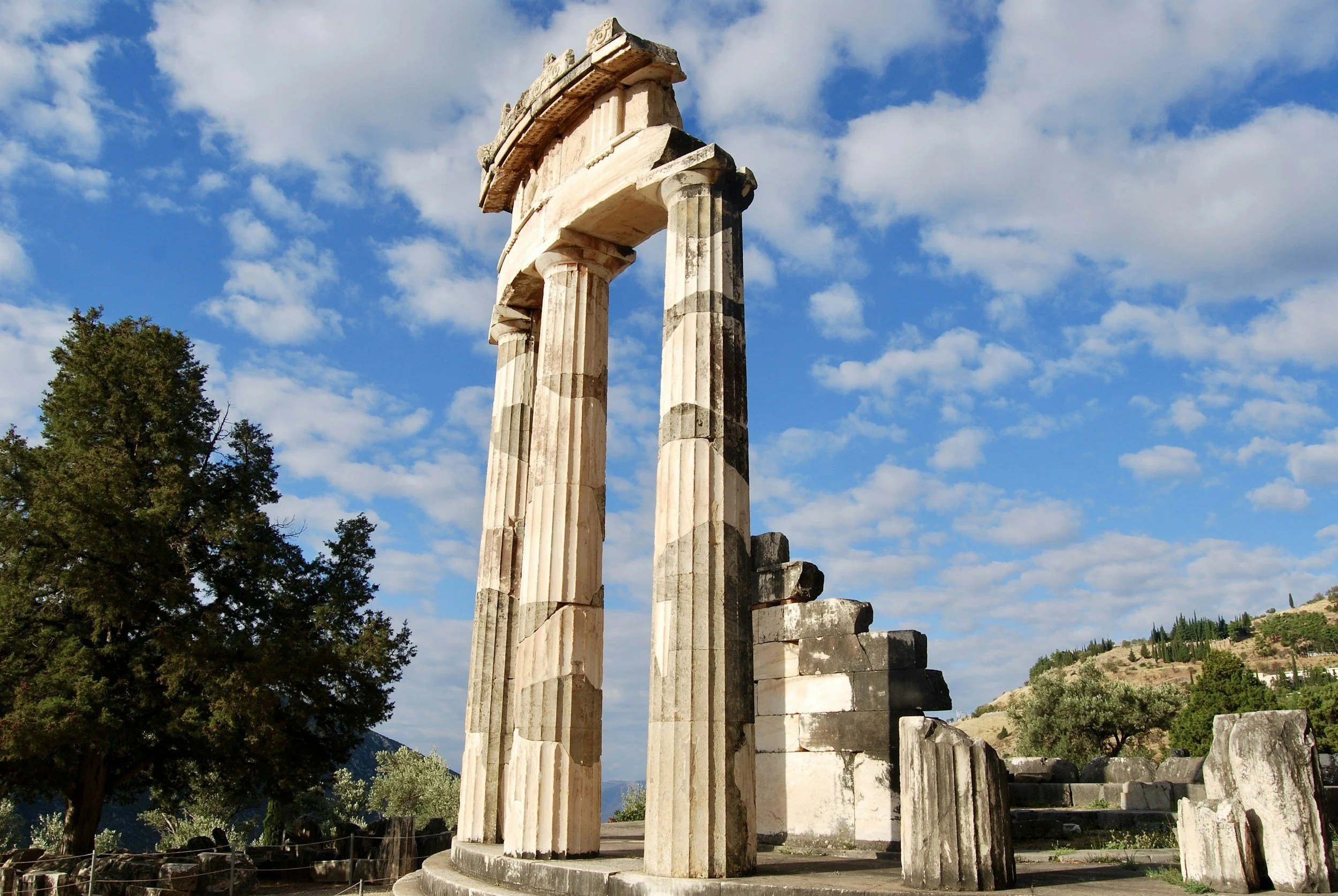 Ancient Greek ruins with tall marble columns against a partly cloudy sky and green landscape in the background.