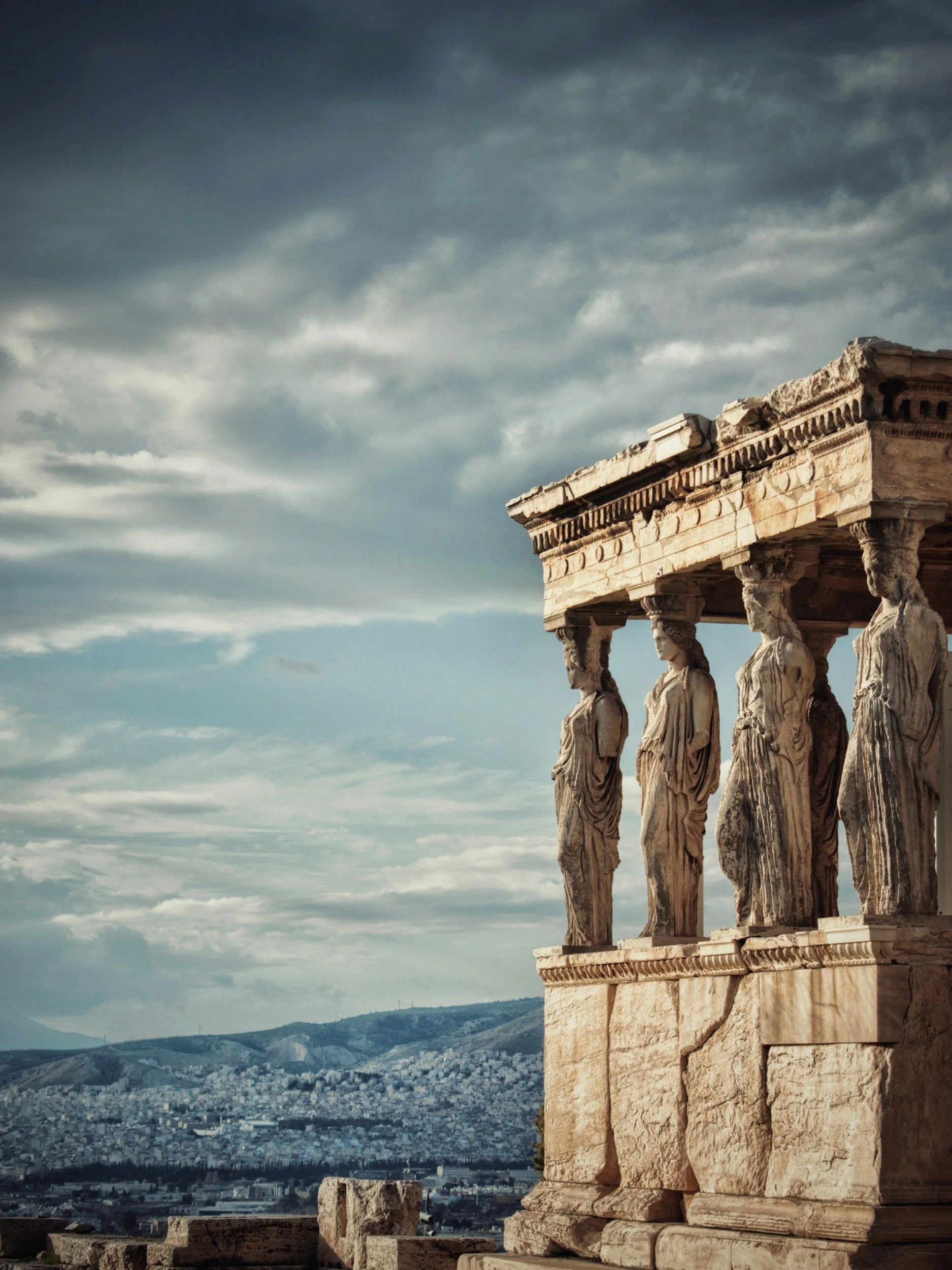 Ancient Greek theater structure with detailed statues of figures supporting the upper platform, set against a cloudy sky and a view of a modern city and mountains in the distance.