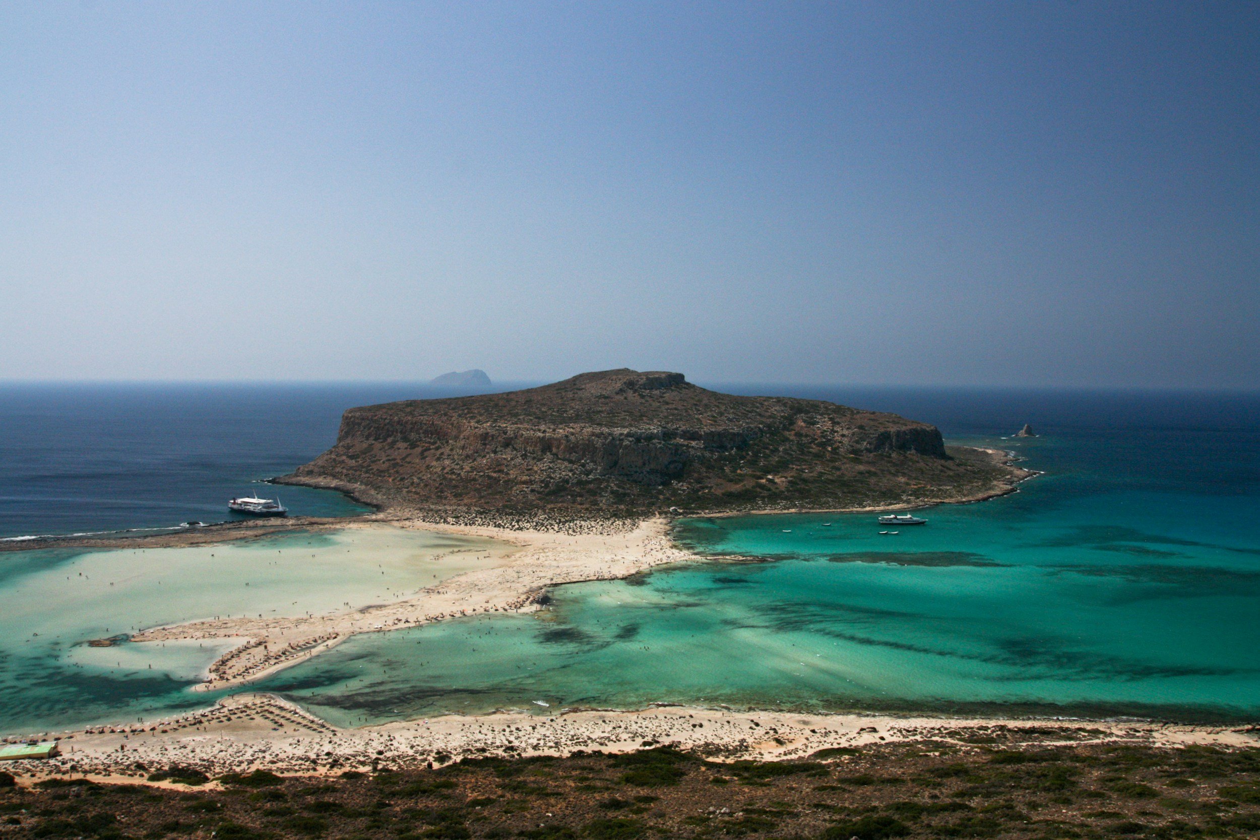 A scenic view of a small island with a flat top and rocky cliffs, surrounded by turquoise water and a white sandy beach, with boats anchored near the shore and a larger ship nearby. The background has a clear blue sky and a distant landmass or island.