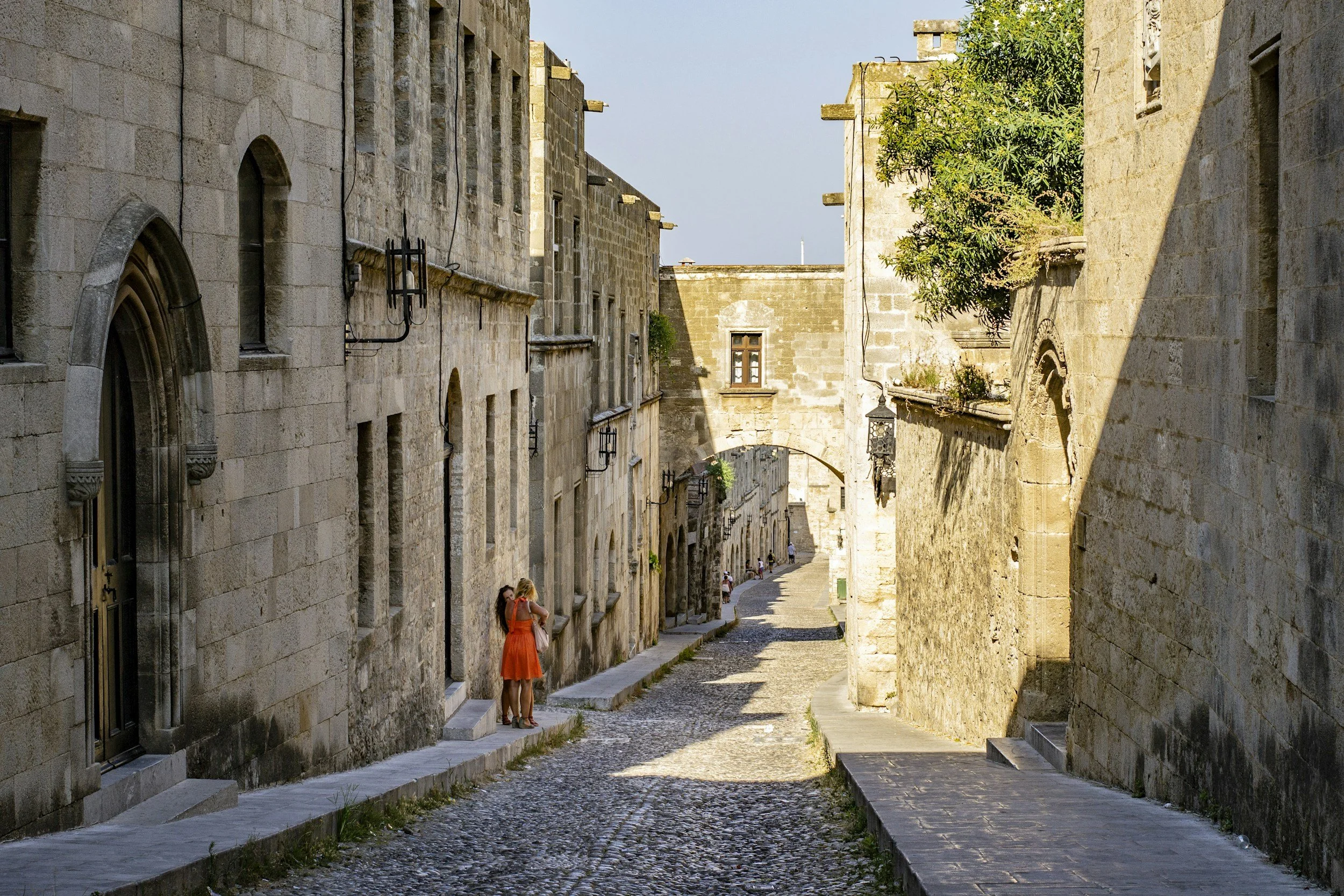 A narrow cobblestone street flanked by old stone buildings, with a few women standing and talking on the sidewalk.