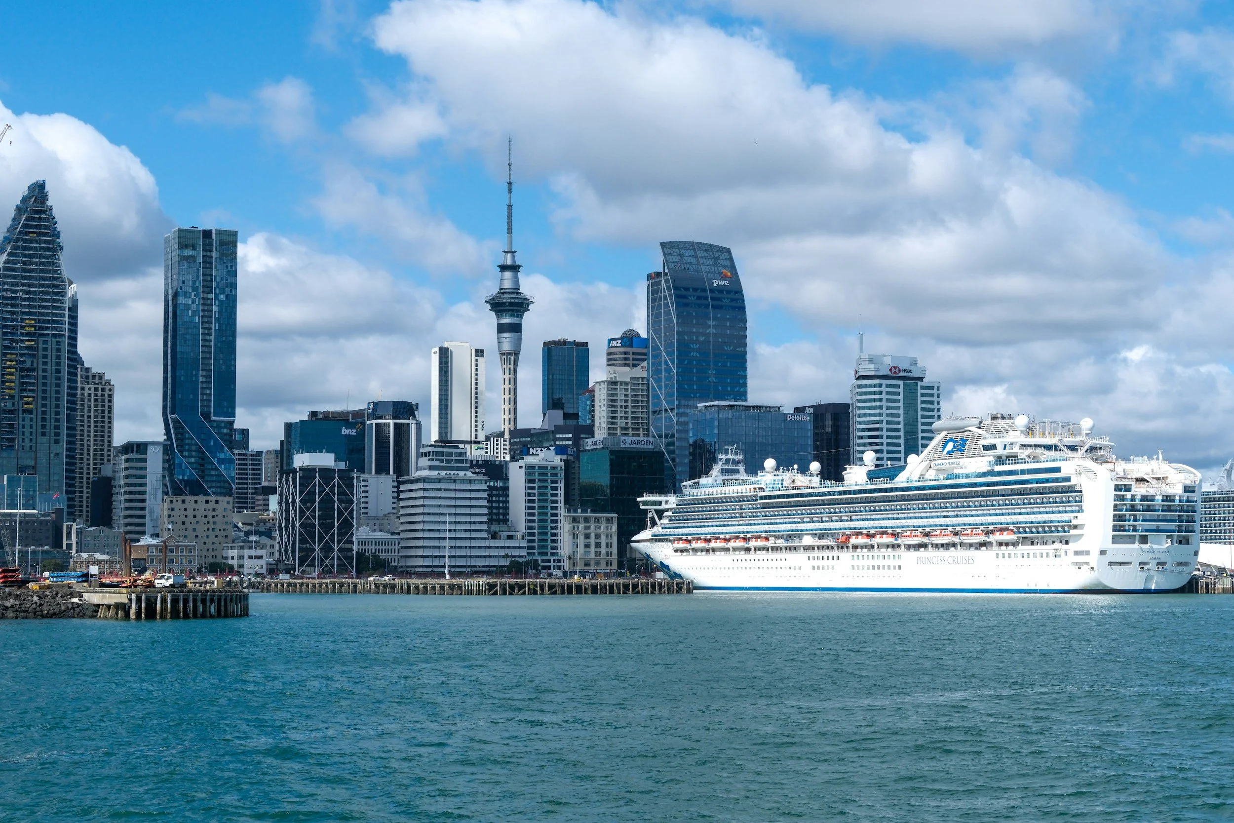 View of a city skyline with tall modern buildings, a tower with an antenna, and a cruise ship docked in the harbor on a partly cloudy day.