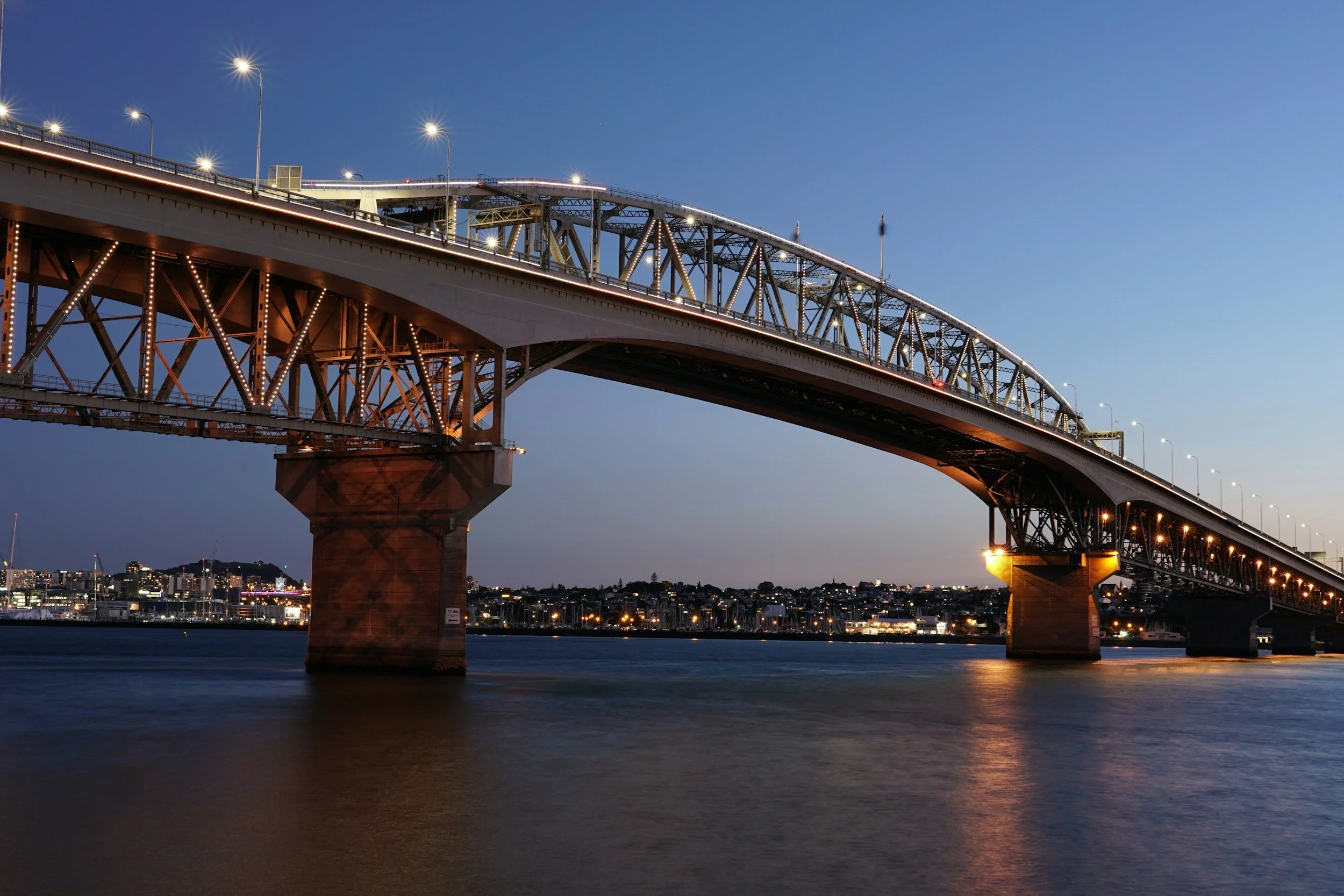 A large steel arch bridge illuminated by lights at dusk, spanning over a body of water with a city skyline in the background.