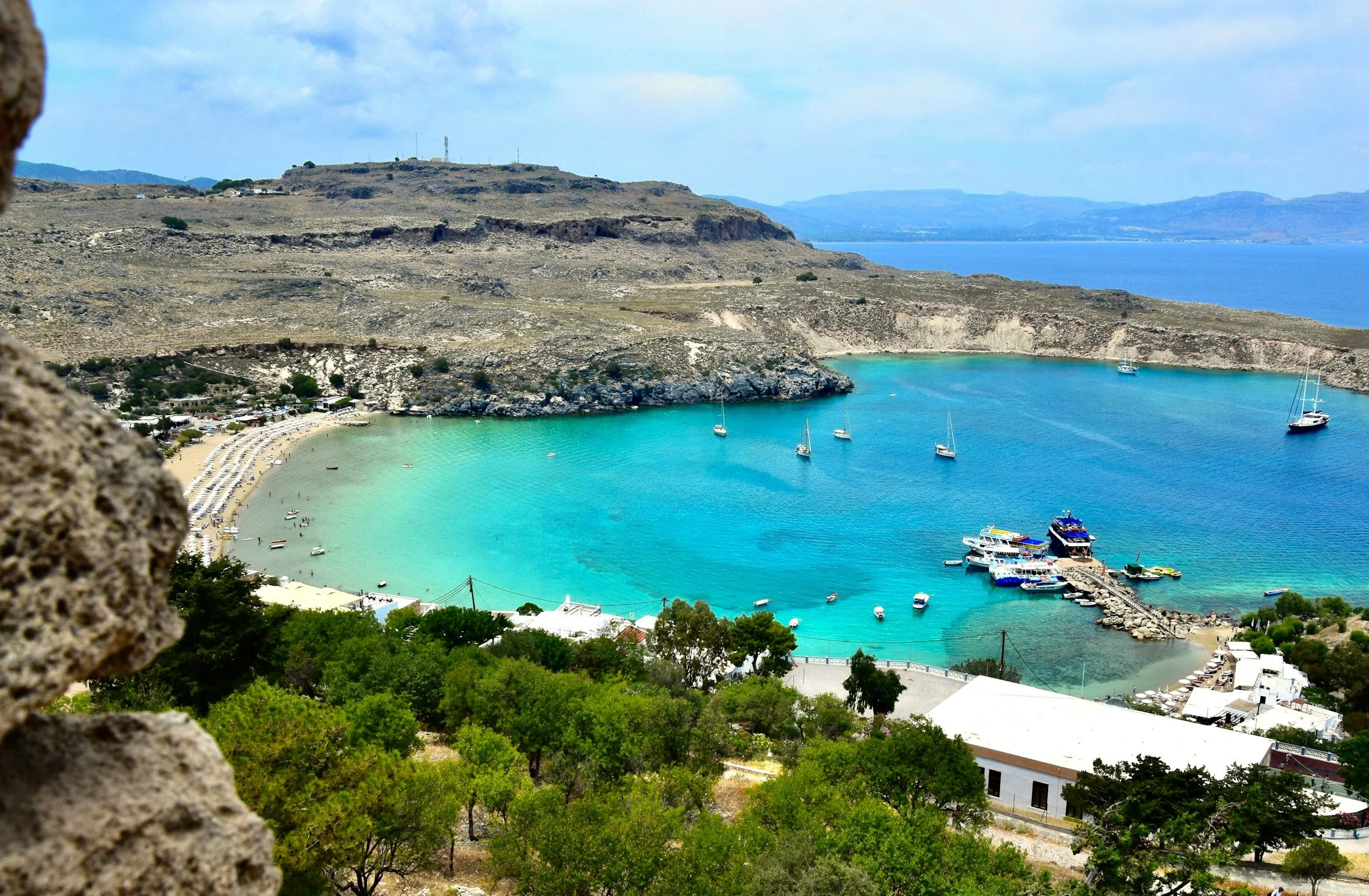 A view overlooking a turquoise bay with boats, surrounded by rocky hills and some buildings, and a partly cloudy sky.