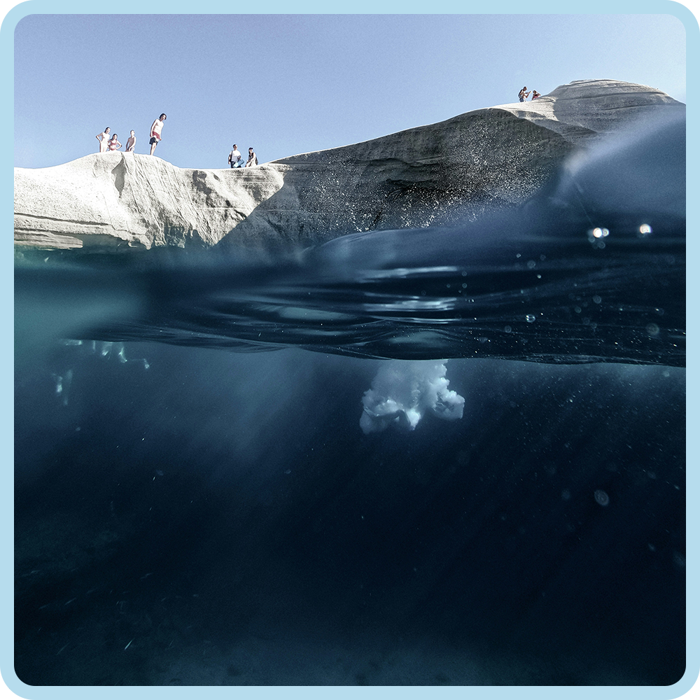 People standing on rocky cliffs at the water's edge, with one person jumping into the water below, and a piece of seafoam or waves beneath the water.