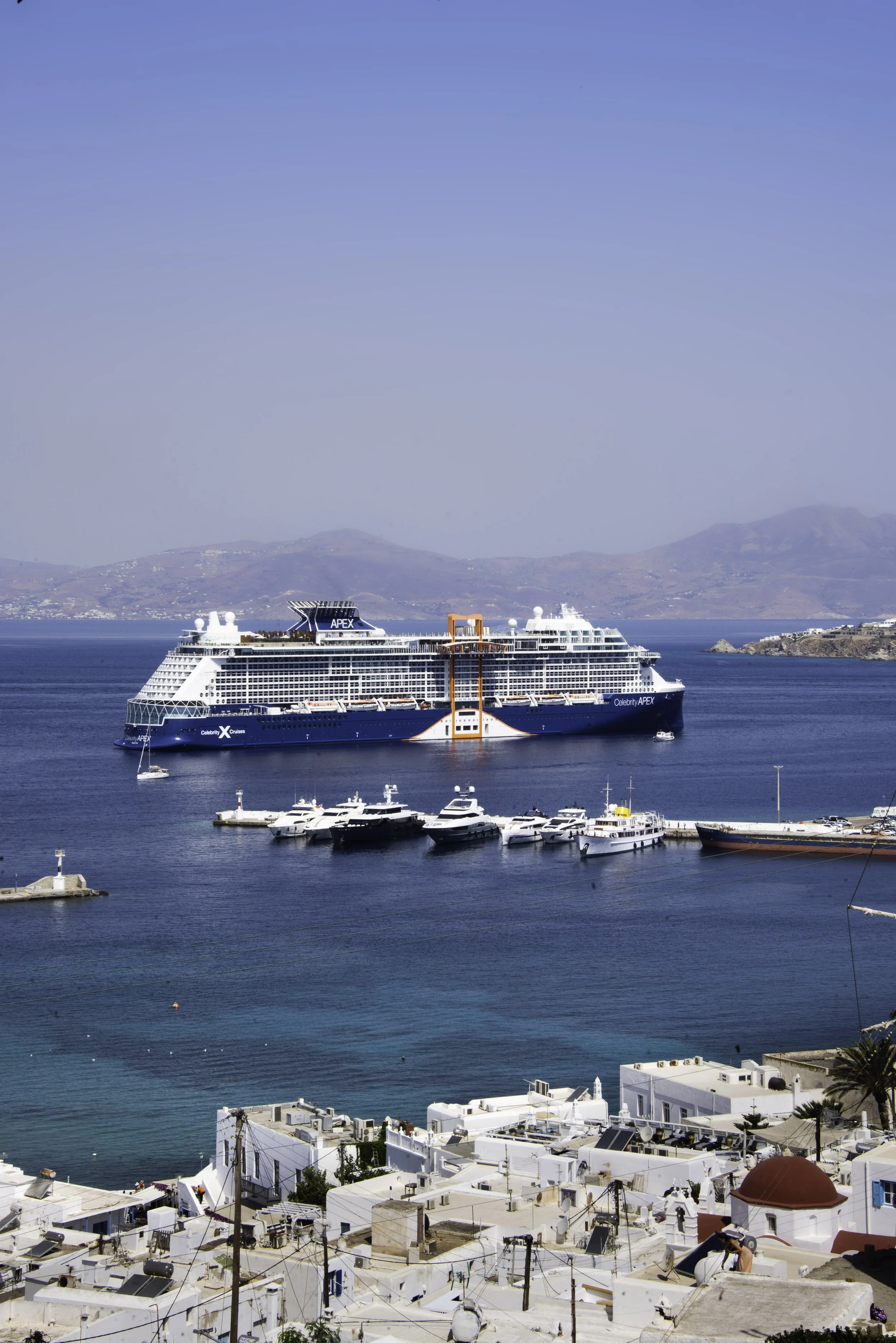 A cruise ship named Celebrity Apex sailing near a coastal town with white buildings and a church with a red dome, against a backdrop of mountains and a clear blue sky.