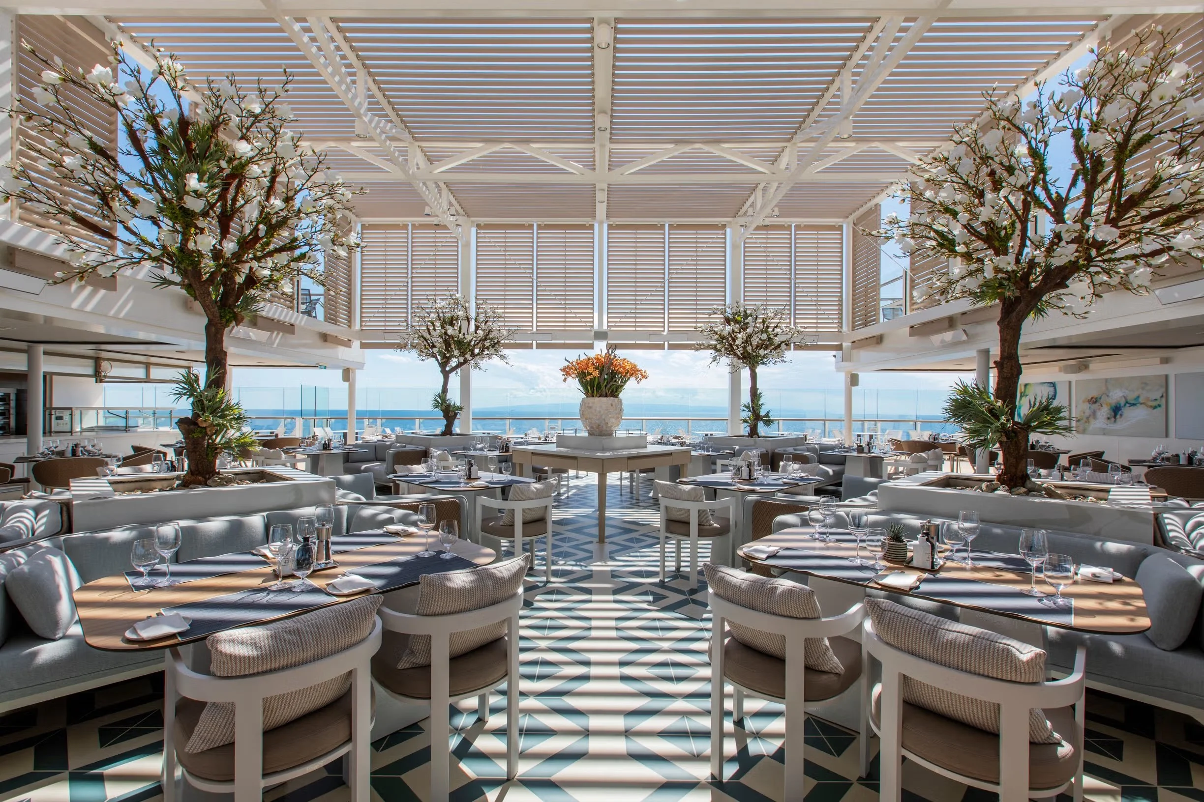 Elegant restaurant with round tables set for dining, decorated with plates, glasses, and napkins. The space features large windows with ocean views, white furniture, and decorative trees with white blossoms. A large central vase with orange flowers is visible, and the ceiling consists of slatted panels allowing natural light.