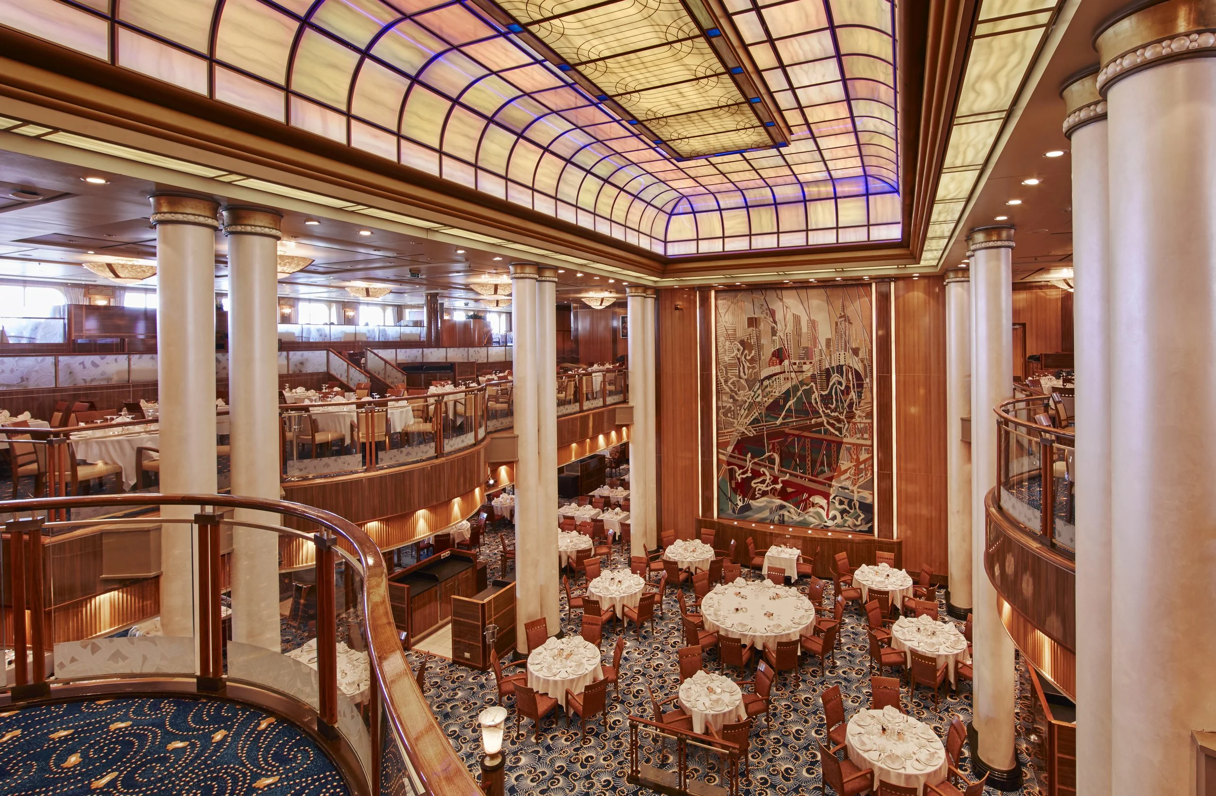Elegant dining room inside a cruise ship with tables set for a meal, decorated with white tablecloths and floral centerpieces. The room features a high, arched ceiling with stained glass panels, large columns, and a decorative carpet with a nautical pattern. The space is well-lit with warm lighting and has an inviting atmosphere.