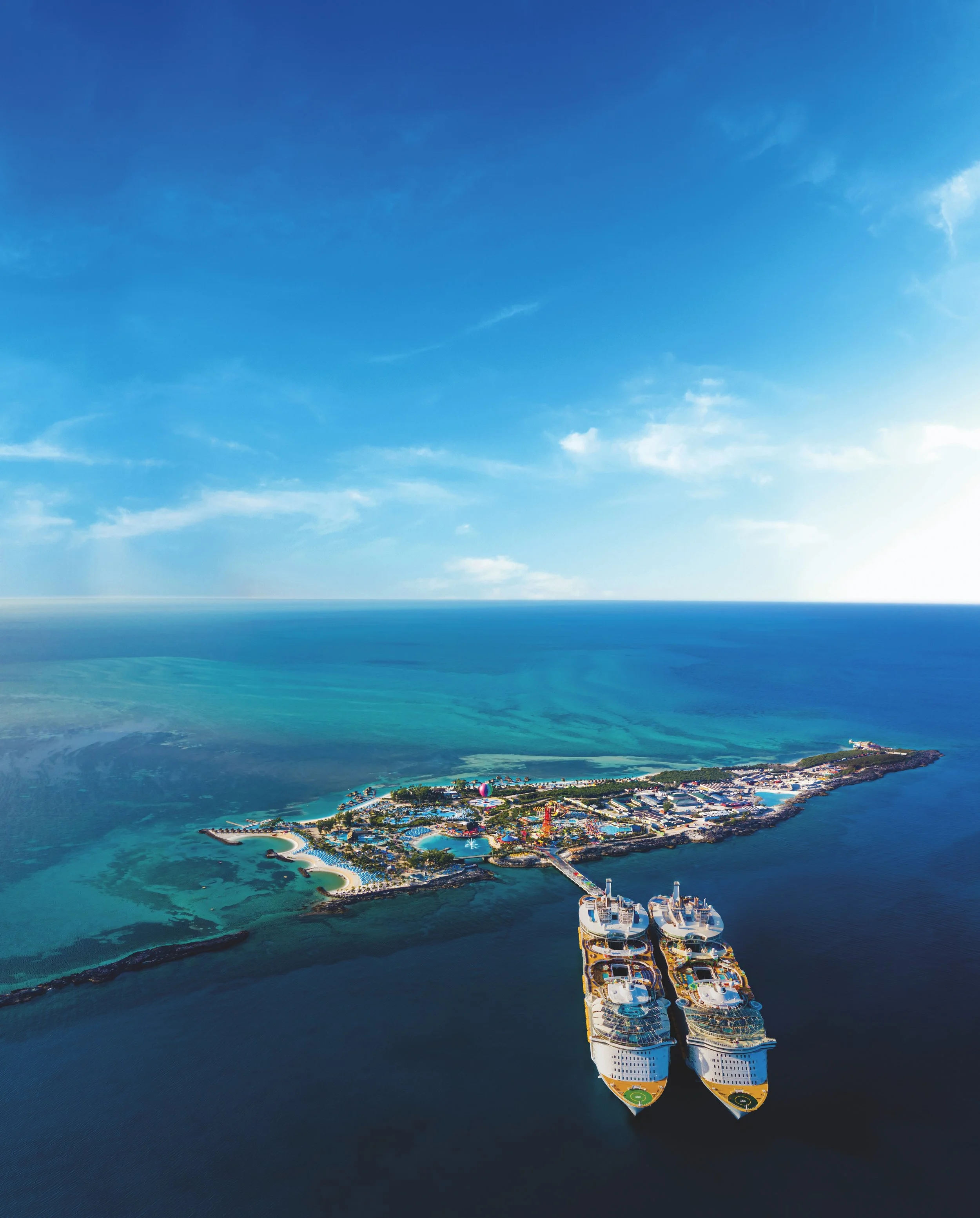 An aerial view of a cruise ship docked at a tropical island with a resort, beaches, and turquoise waters.