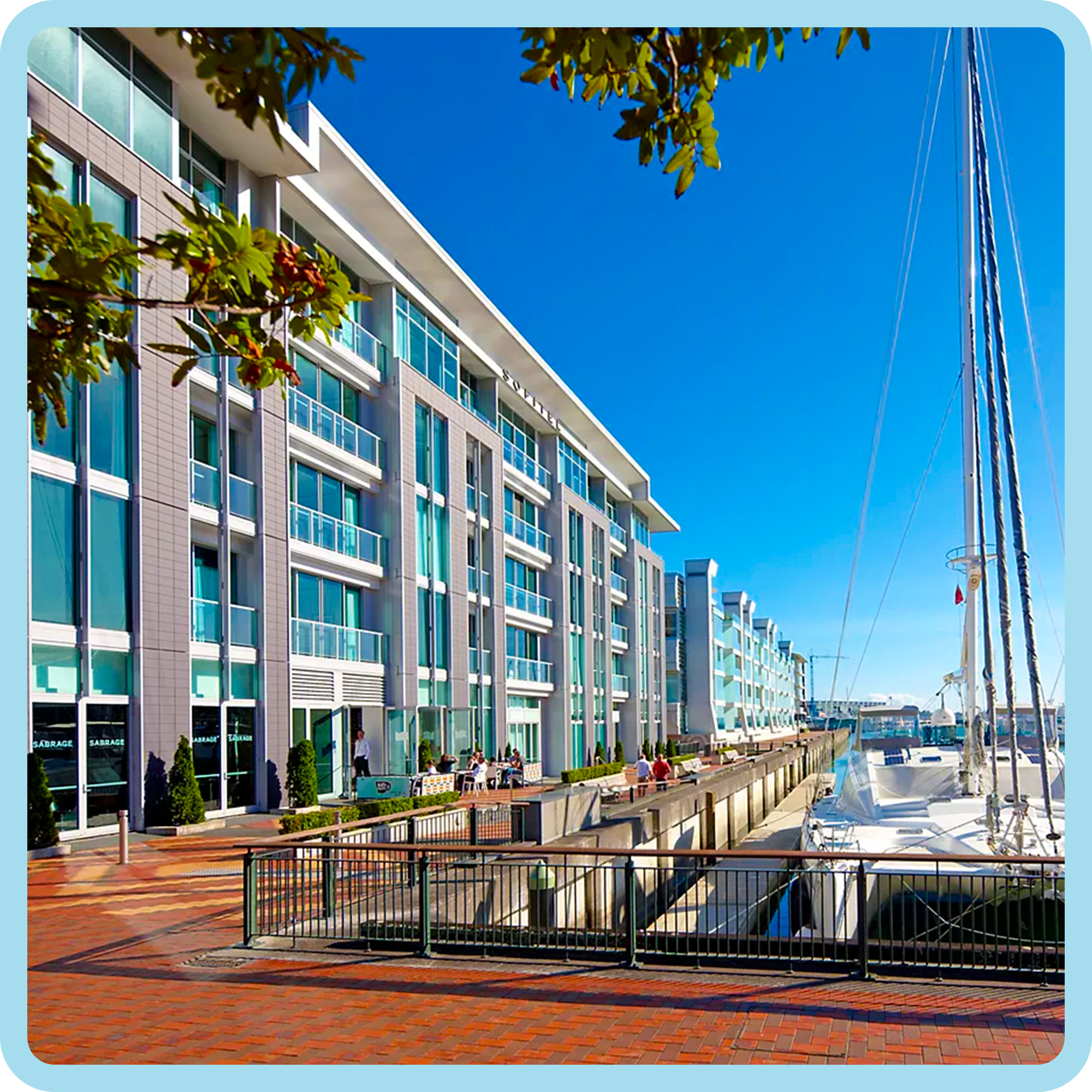 A marina with boats docked, a pathway with a brick pattern, a modern multi-story building with large glass windows, and trees under a bright blue sky.