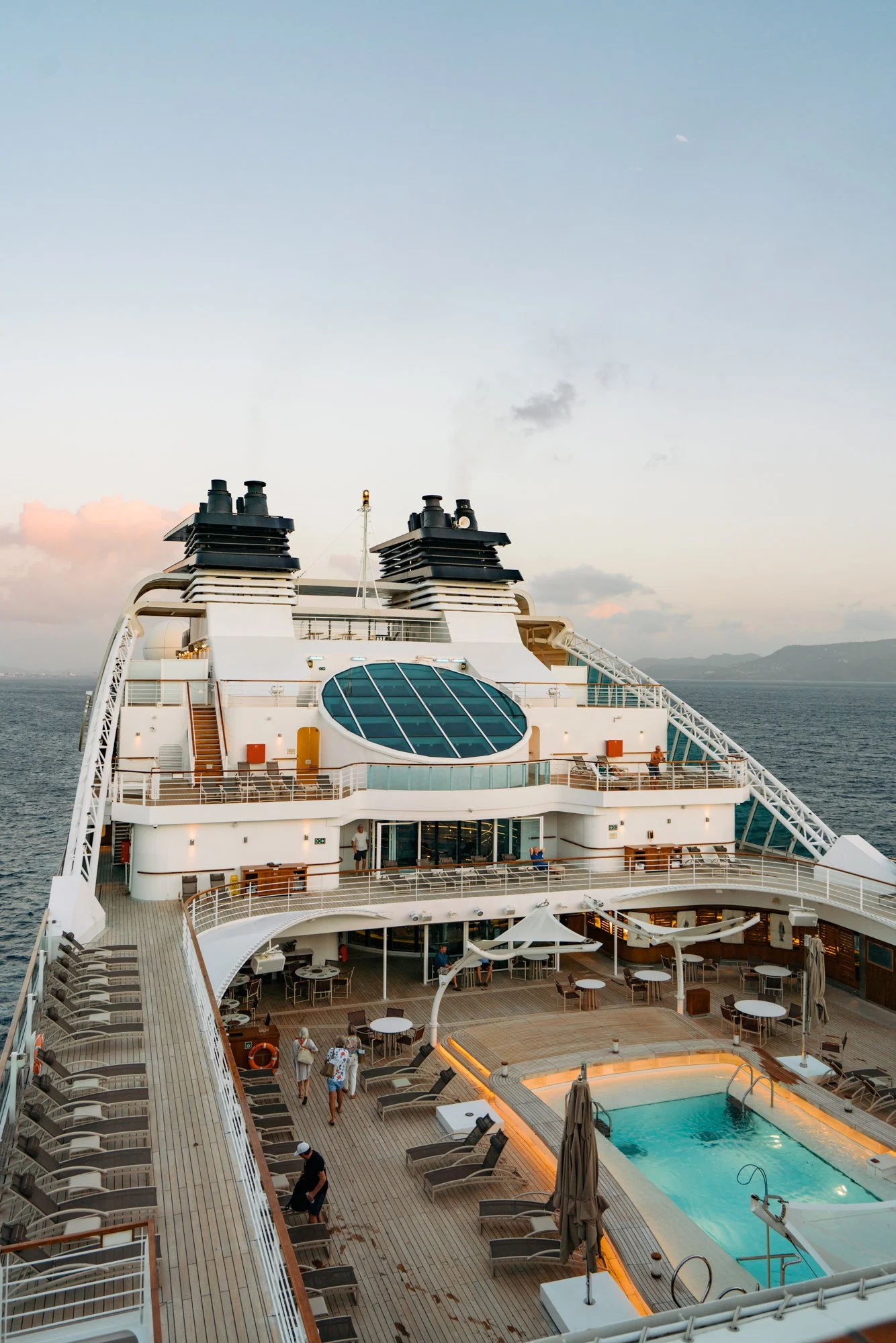 View of the upper deck of a cruise ship with outdoor seating, a swimming pool, and people relaxing during sunset, with the ocean and distant land in the background.