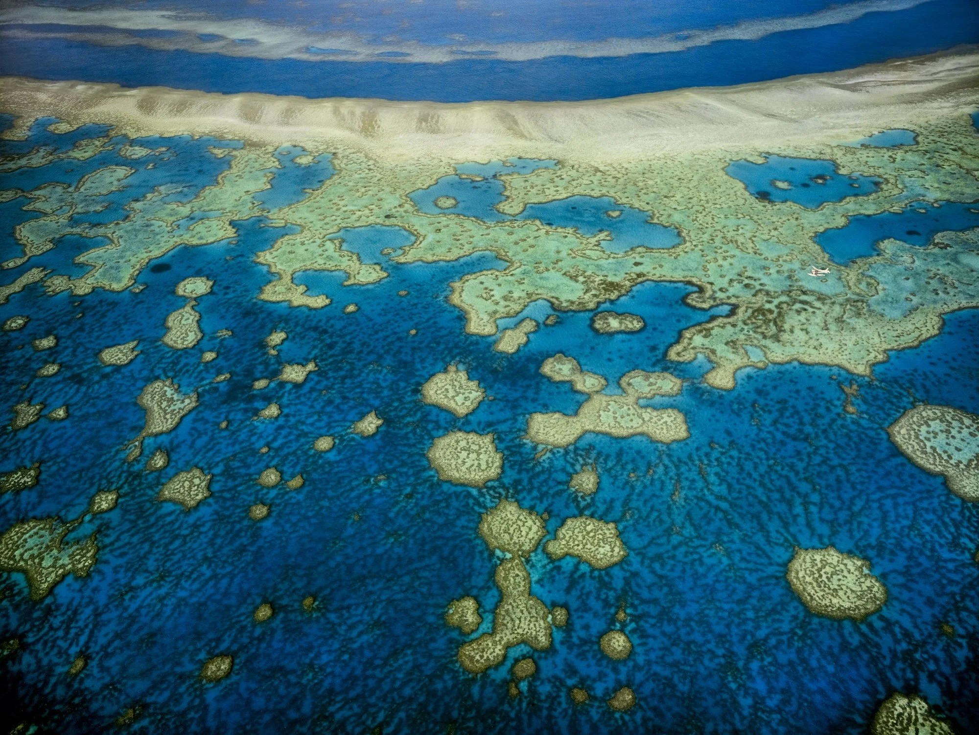 A satellite view of a tropical coral reef with clear blue waters and small islands, with a small airplane flying above the water.