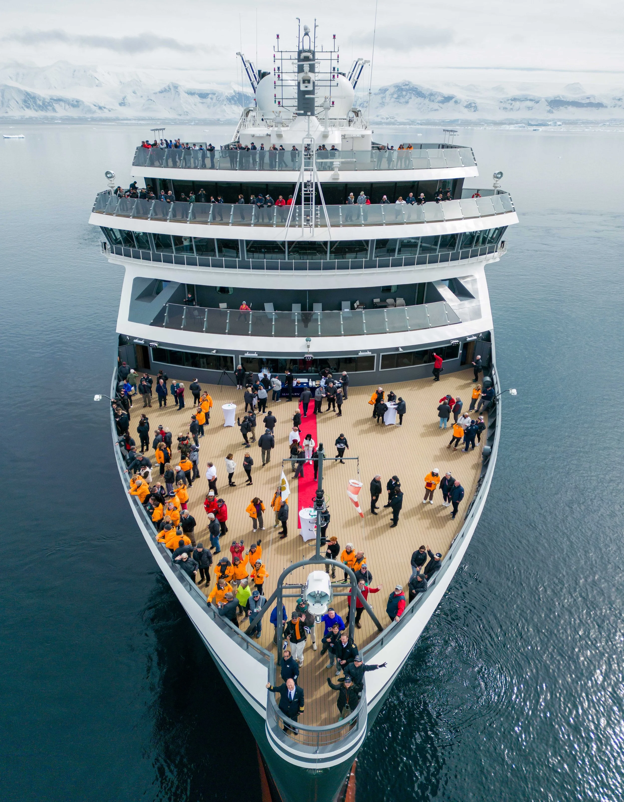 A large cruise ship on icy waters with many passengers on the decks, set against a backdrop of snow-covered mountains and cloudy sky.