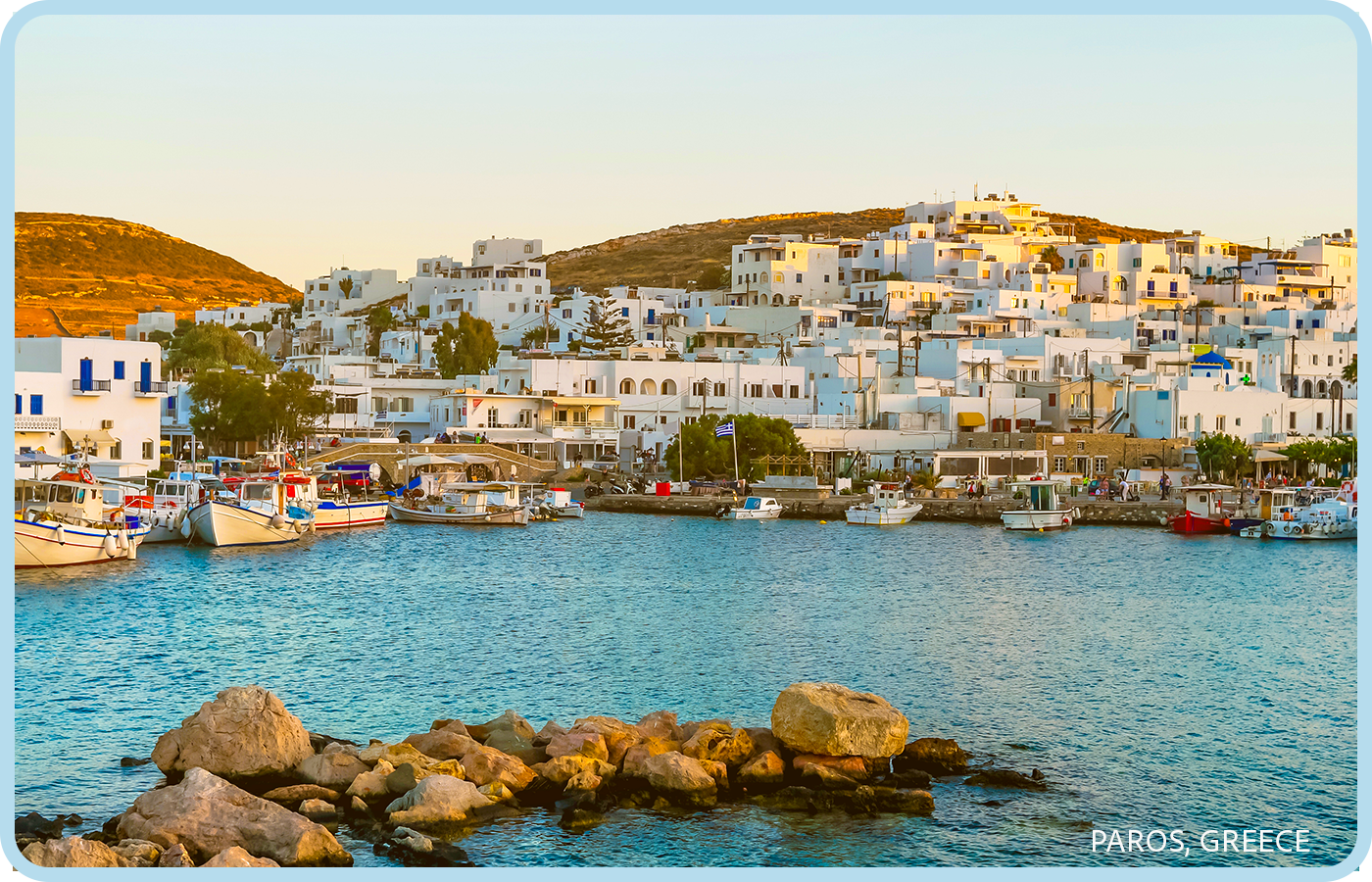 A scenic view of the harbor in Paros, Greece, with white buildings on hilly terrain, boats docked along the shoreline, and calm water in the foreground.