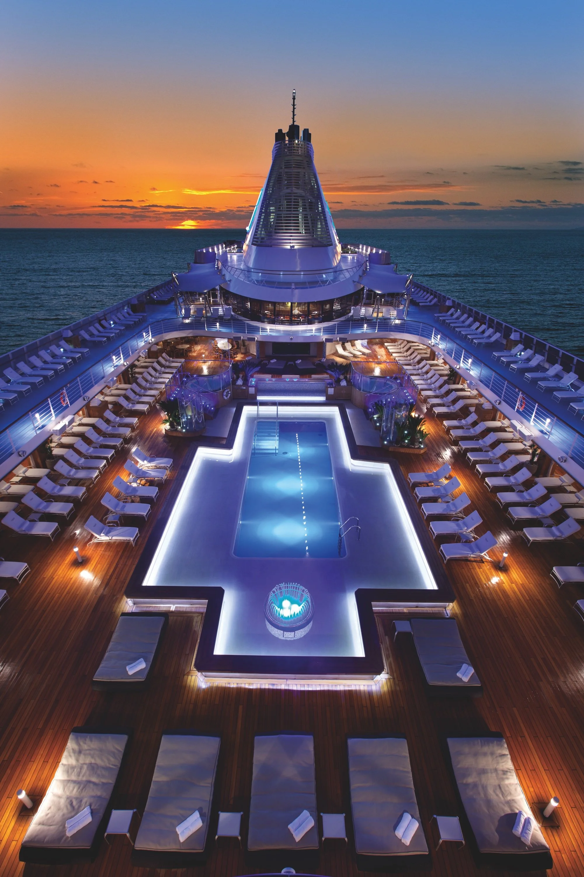 A luxury cruise ship deck at sunset with a rectangular swimming pool surrounded by lounge chairs and shaded areas, overlooking the ocean.