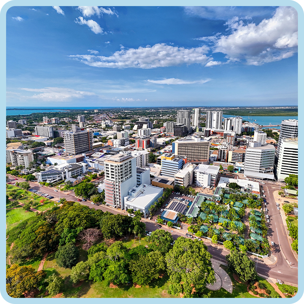 Aerial view of a cityscape with modern high-rise buildings, a park with lush green trees, and a body of water in the background under a partly cloudy sky.