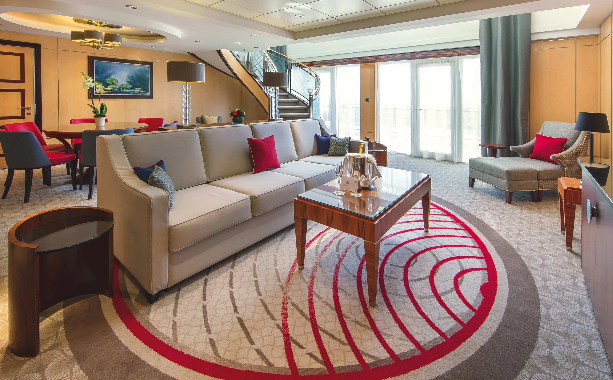 Luxurious hotel living room with cream sofa, covered coffee table, and colorful throw pillows. A round wooden dining table with red and gray chairs is alongside. Large windows with sheer curtains let in natural light. Decor includes two modern lamps, a painting of water lilies, and a staircase with metal and glass railing. An elegant patterned rug features red and gray concentric circles.