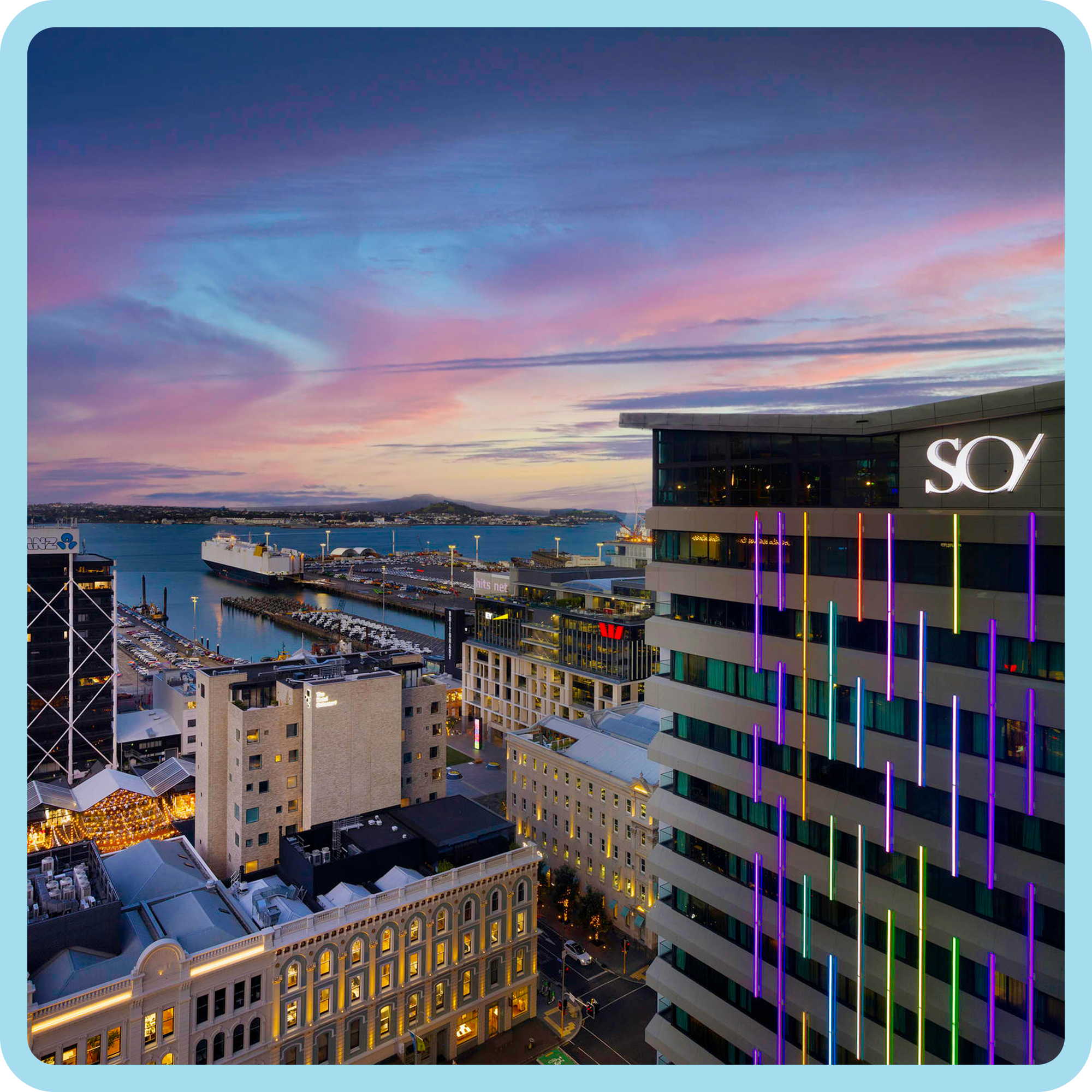 View of a harbor with ships and buildings at sunset, with a colorful sky and cityscape in the background.