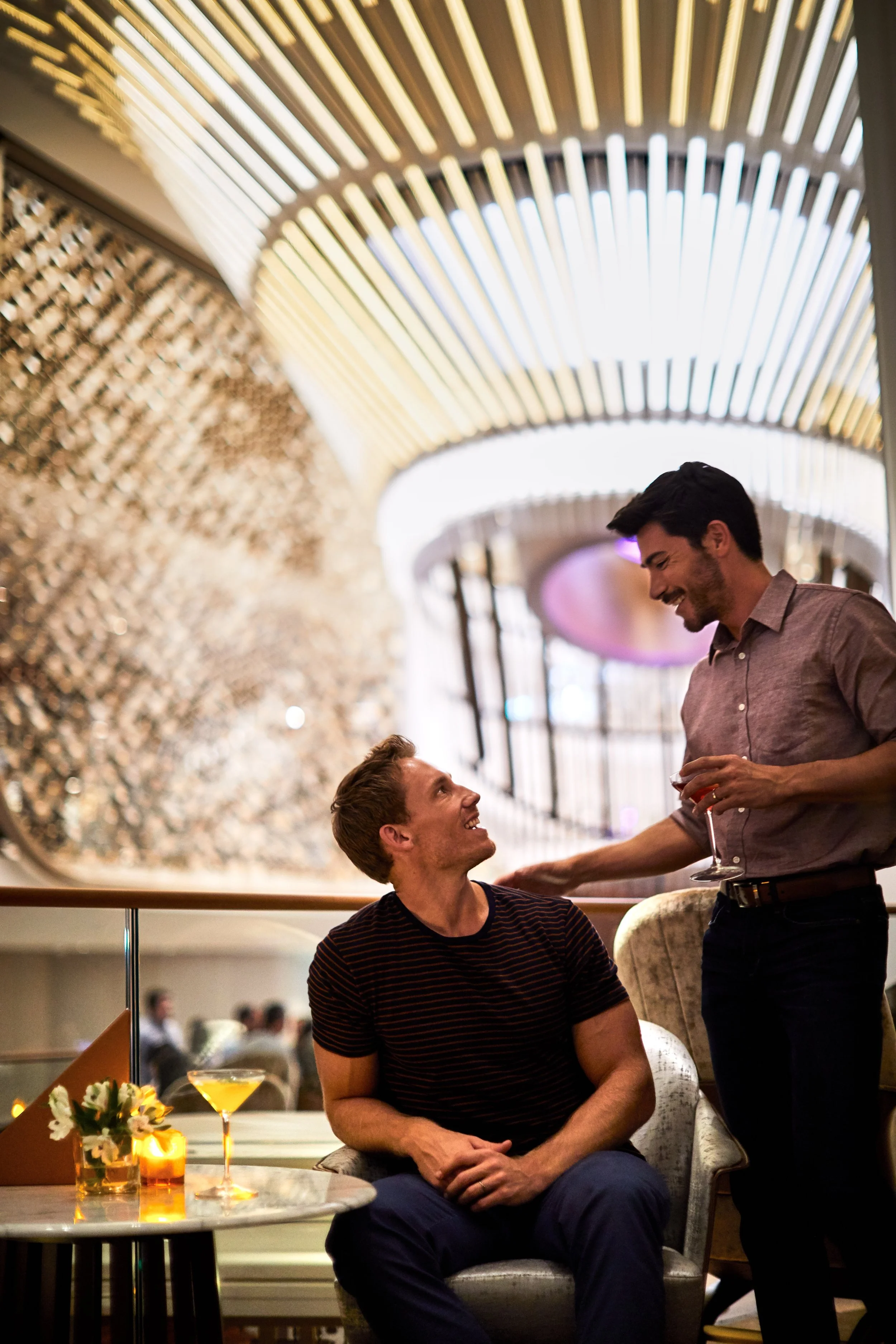 Two men are in a lively restaurant or bar. One man, sitting at a table with a cocktail, is looking up and smiling at the standing man. The standing man is holding a glass and smiling back. The setting is modern and stylish with decorative ceiling panels.