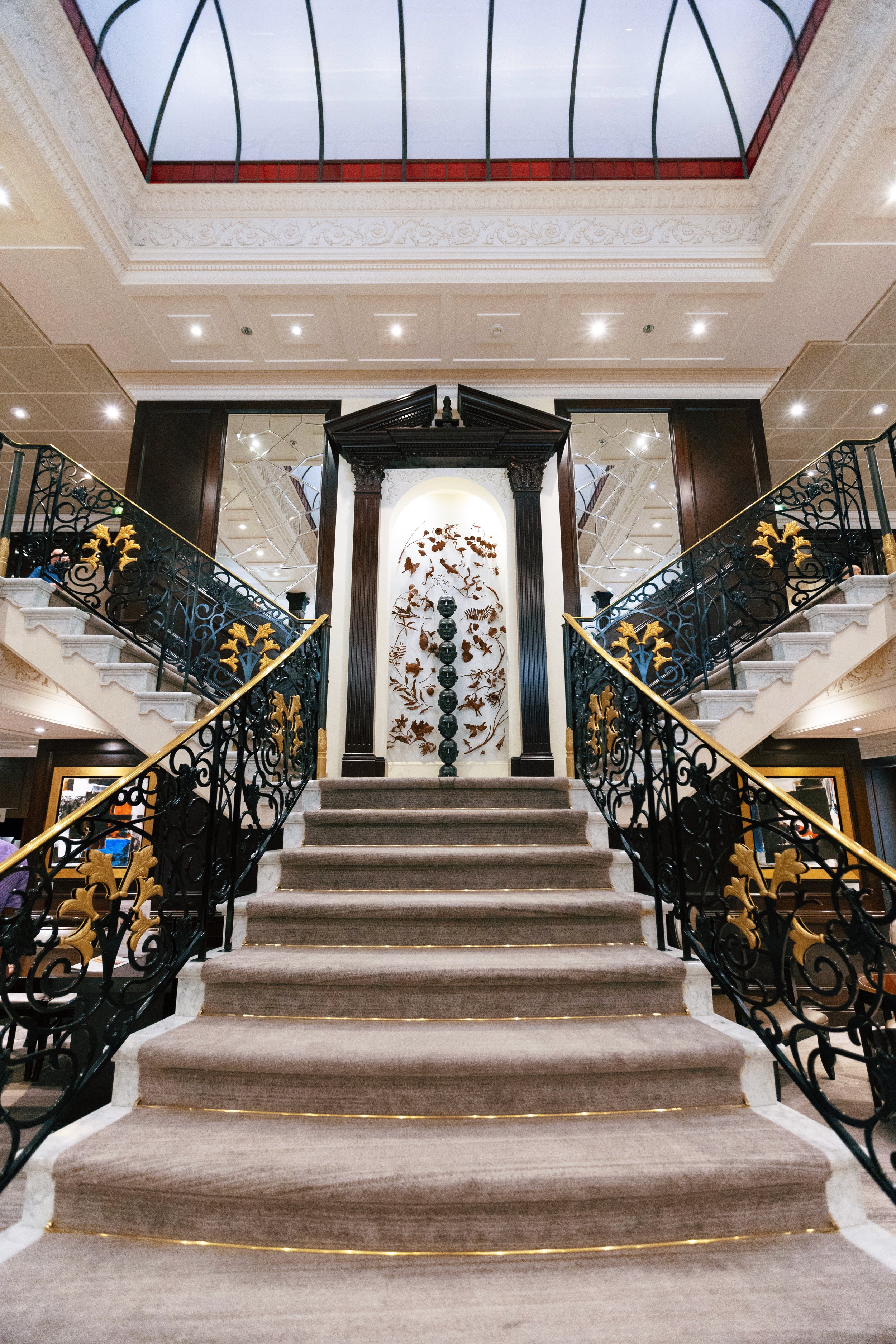 Elegant staircase with ornate black and gold wrought iron railings, gray carpeted steps, and a decorative art piece with human heads and botanical motifs in a luxurious interior.