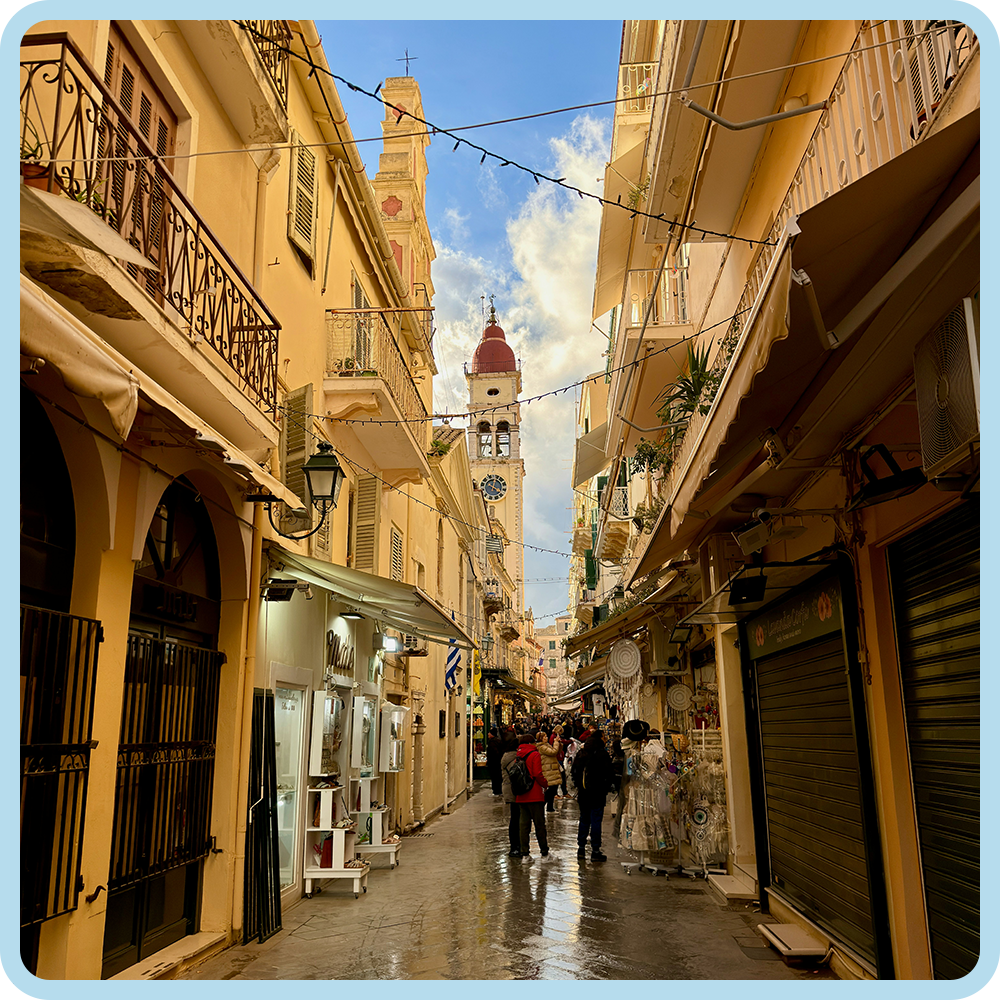 A narrow street in a European city with yellow buildings, balconies, shops, and a clock tower in the background, during daytime with a partly cloudy sky.
