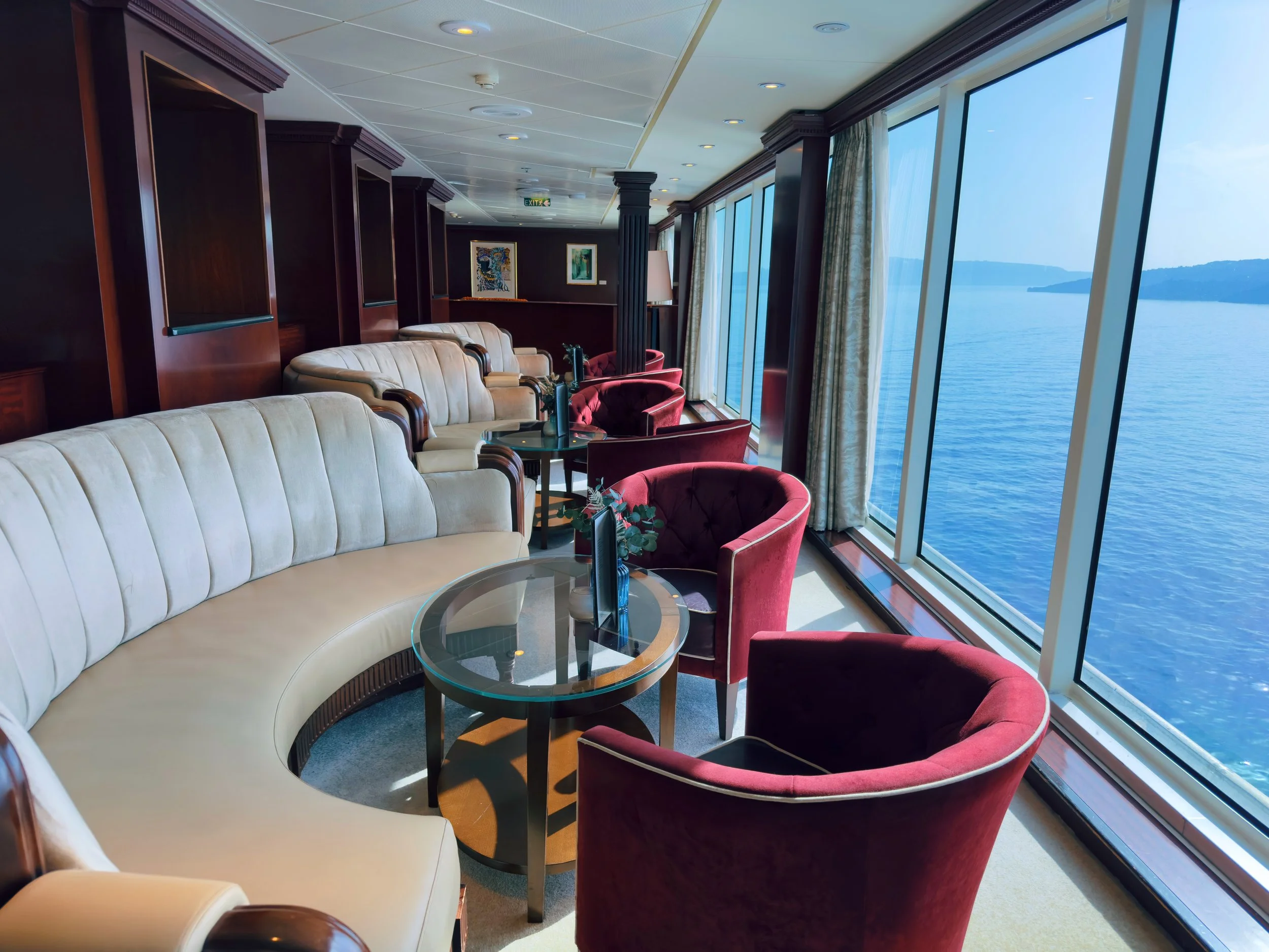 Interior of a lounge or sitting area on a cruise ship with large windows showing a view of the ocean and distant islands, featuring plush chairs and a glass-top table.