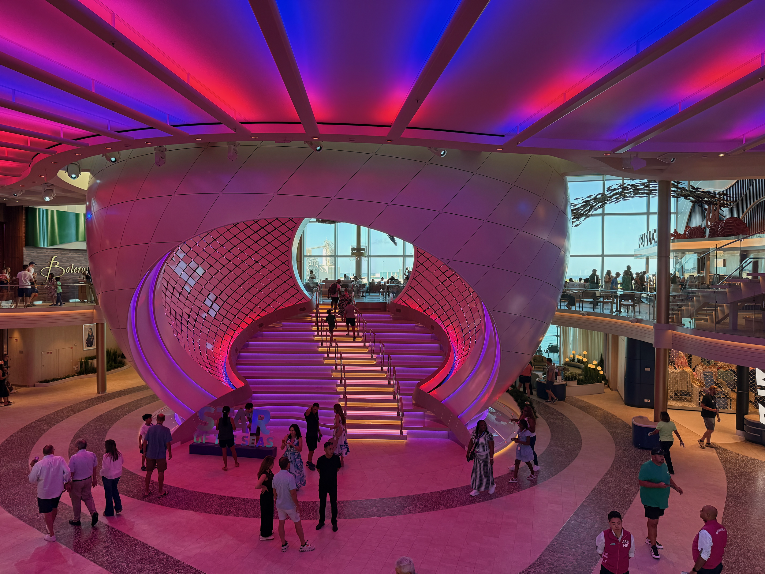Interior view of a Star of the Seas Pearl with a staircase under colorful purple and pink lighting, people walking and sitting, large windows in the background.